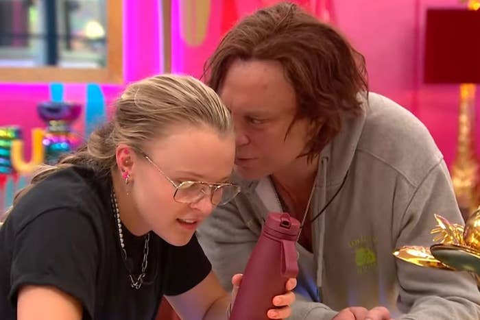 Two individuals are engaged in a conversation. One is wearing glasses and holding a water bottle, and the other is listening closely