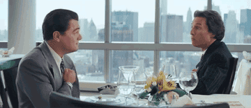 Two men in suits sit at a fancy restaurant table, animatedly talking and gesturing, with a city skyline visible through the large windows behind them