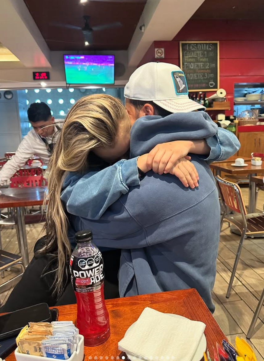 Two people hugging closely at a dining area, one wearing a baseball cap and casual clothing, the other with long hair