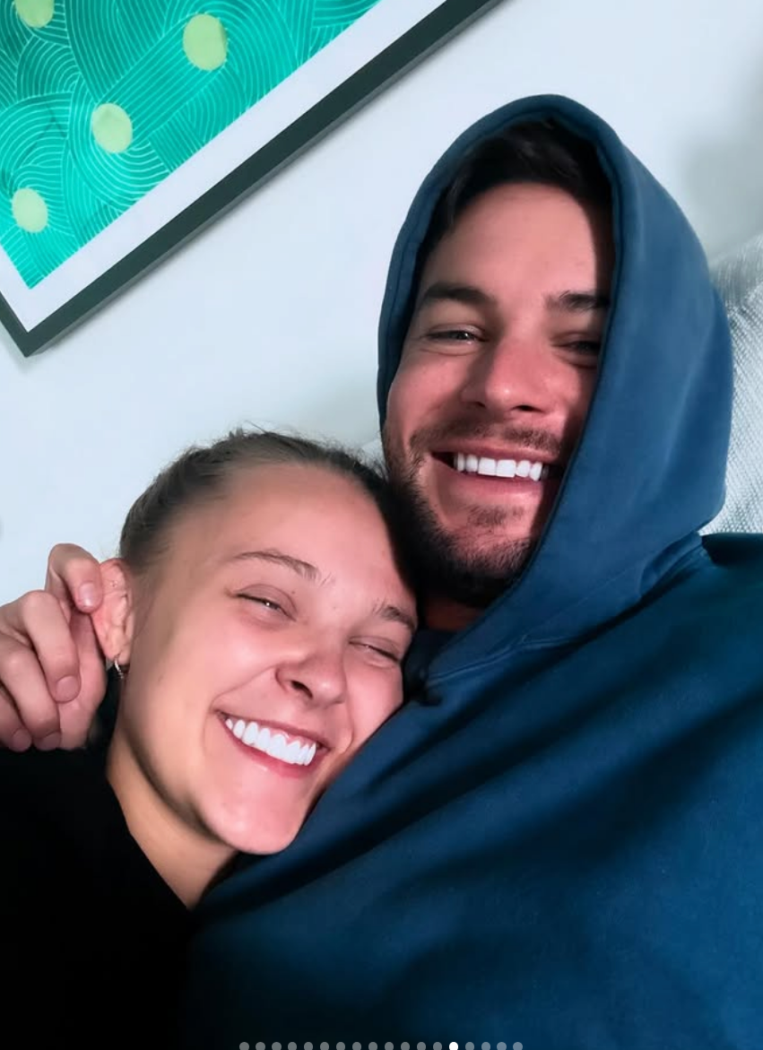 Two people are smiling warmly, snuggled together in casual clothing, with one wearing a hoodie