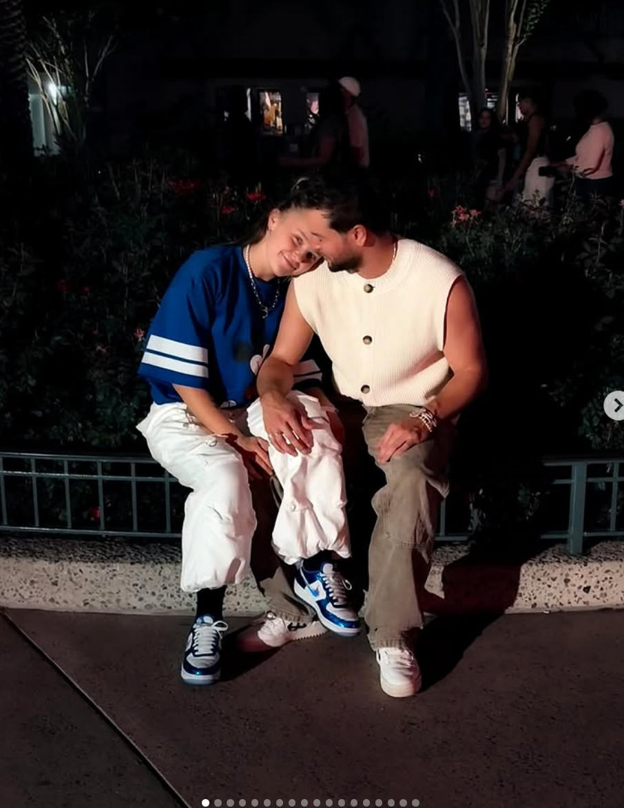 Two people sit together on a bench at night, smiling and leaning toward each other. One wears a blue shirt, the other a sleeveless top