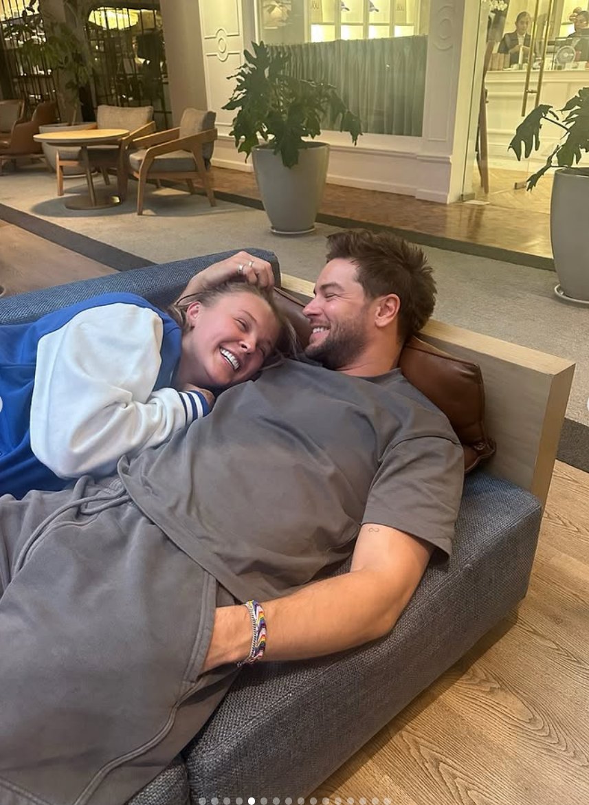 Two people relax and smile on a couch indoors. One wears a varsity jacket, the other a casual t-shirt and shorts