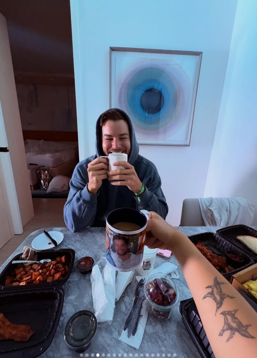 Person in hoodie smiles over coffee at a table with takeout food. Another person holds a mug in the foreground. Room has modern decor