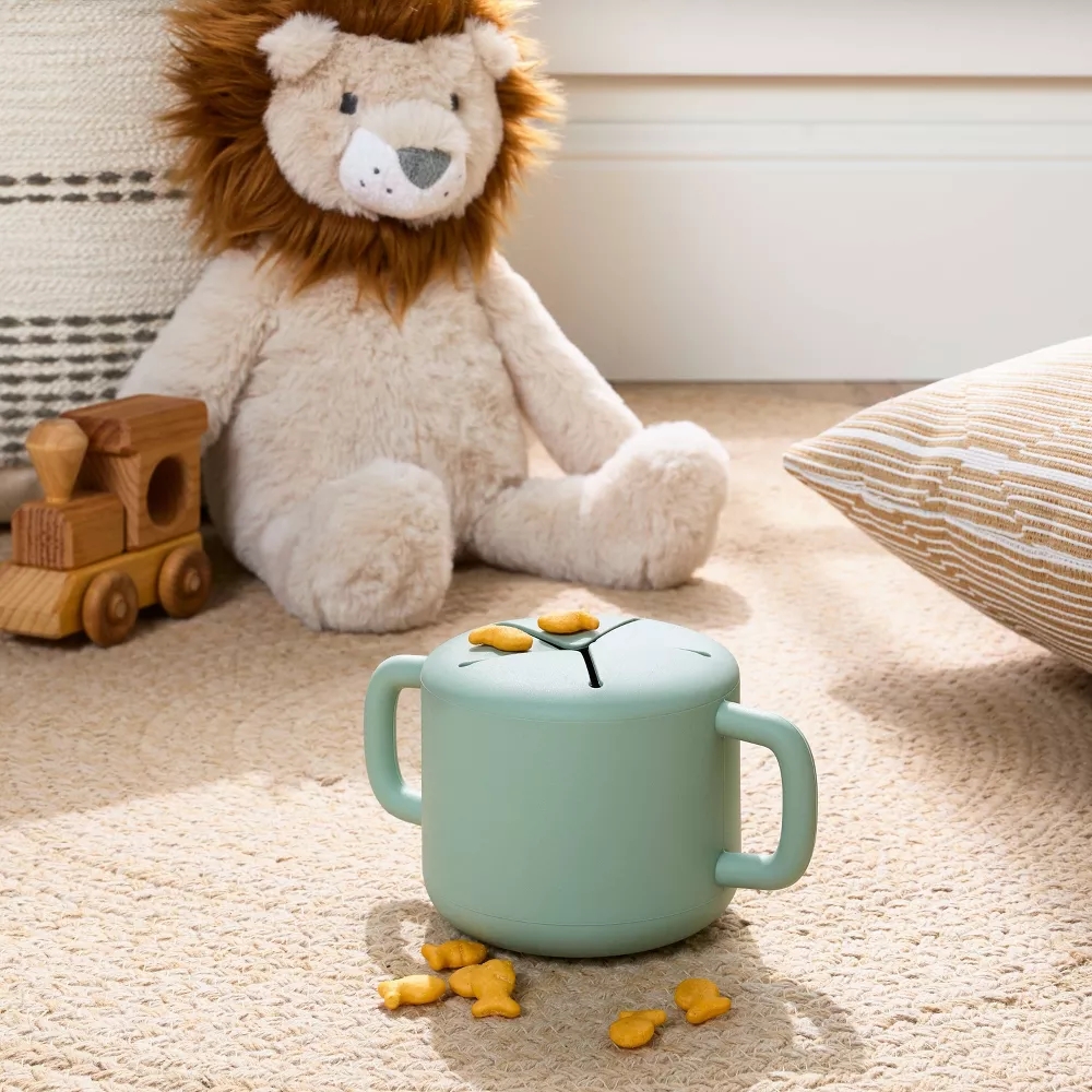 Plush lion toy sits on a carpet beside a wooden toy train, with a snack cup holding fish-shaped crackers nearby