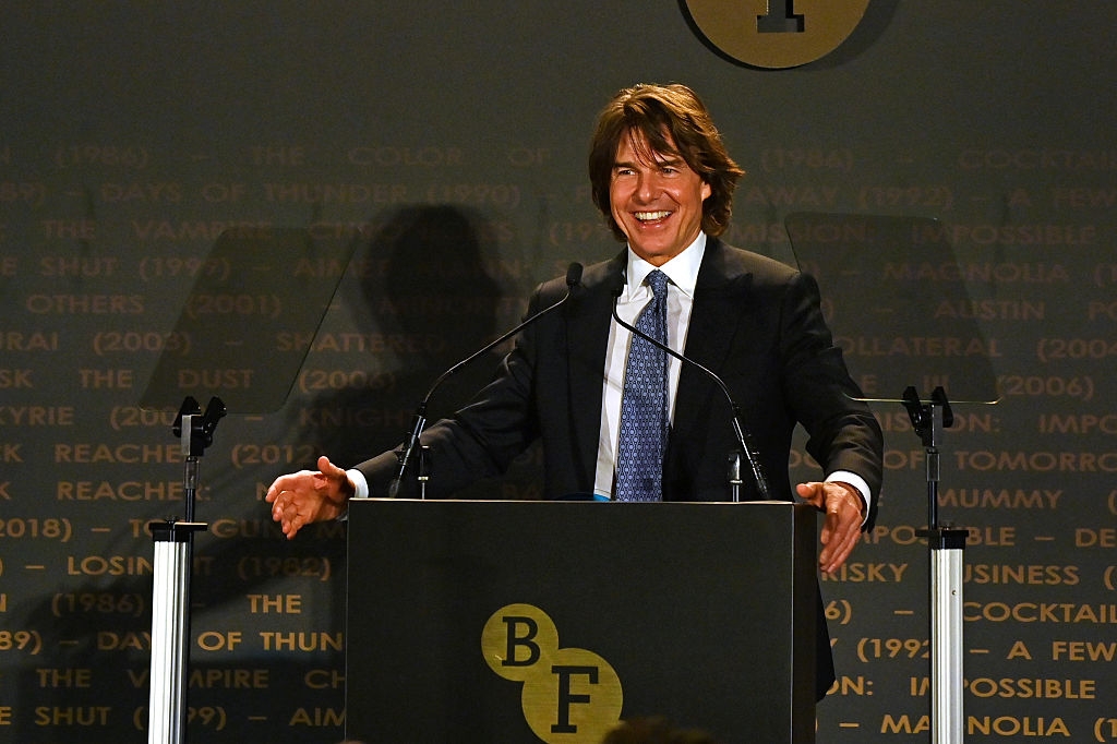 A person in a suit and tie stands at a podium with microphones, smiling, at a formal event backdrop