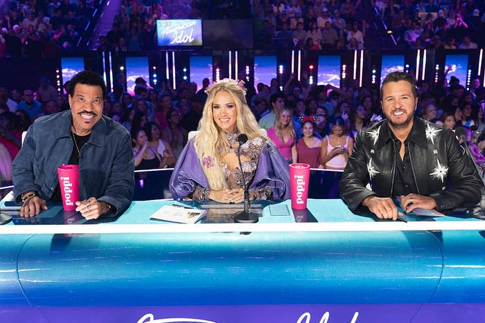 Three talent show judges sit at a panel with audience behind them. One wears a studded leather jacket, the second a gown with embellishments, and the third a denim jacket