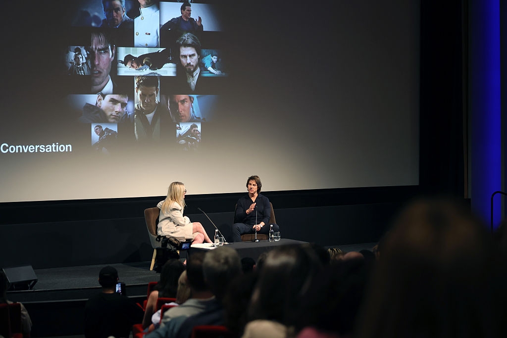 Two people seated on stage discussing in front of a large screen displaying movie scenes. Audience is watching