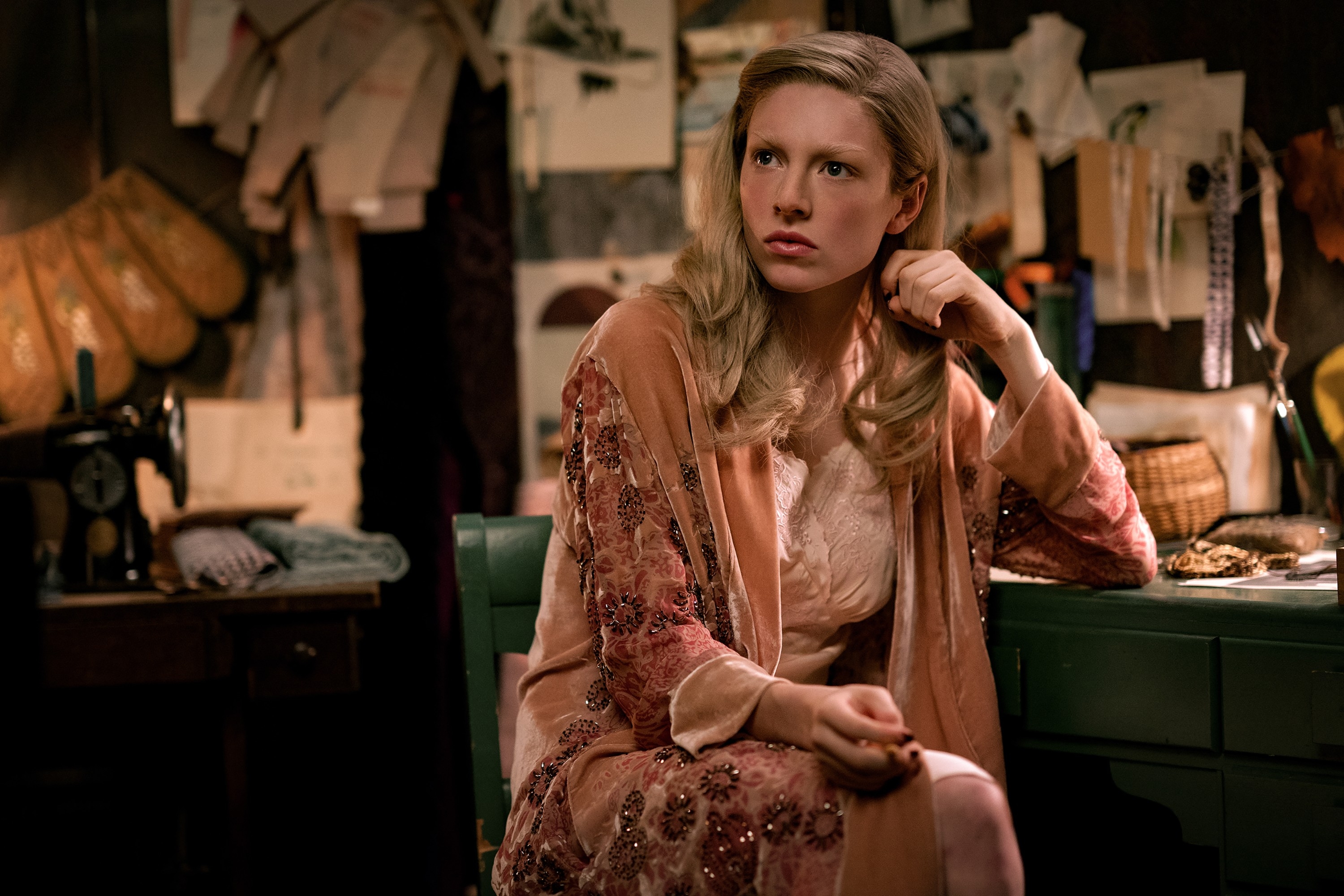 Hunter in vintage-style clothing sits by a sewing machine in a cluttered room, looking contemplative