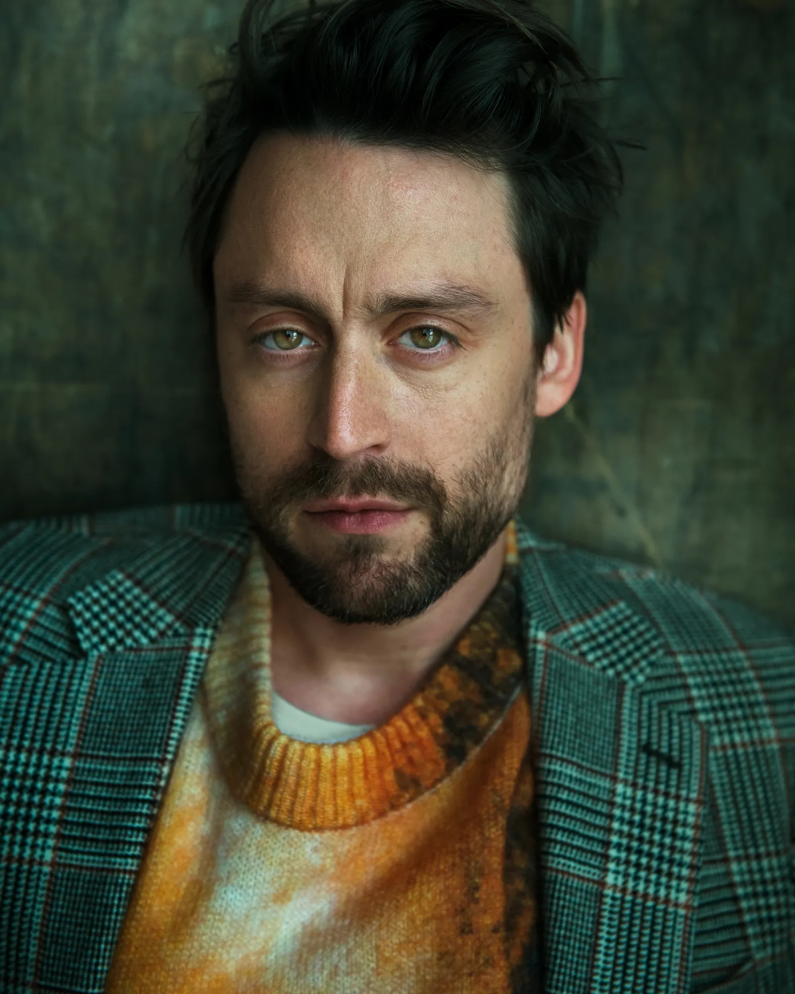 Persona con barba y cabello oscuro viste un suéter estampado debajo de un saco de cuadros. Mira directamente a la cámara