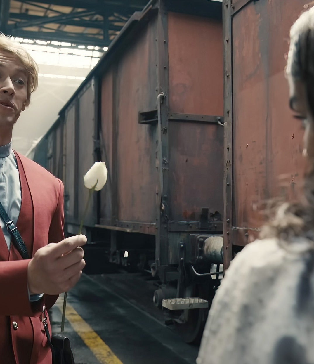 Tom coat holds a flower, near industrial train cars in a warehouse setting