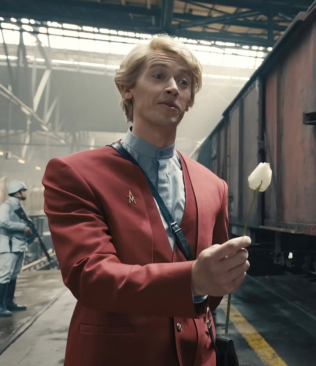 Tom coat holds a flower, near industrial train cars in a warehouse setting