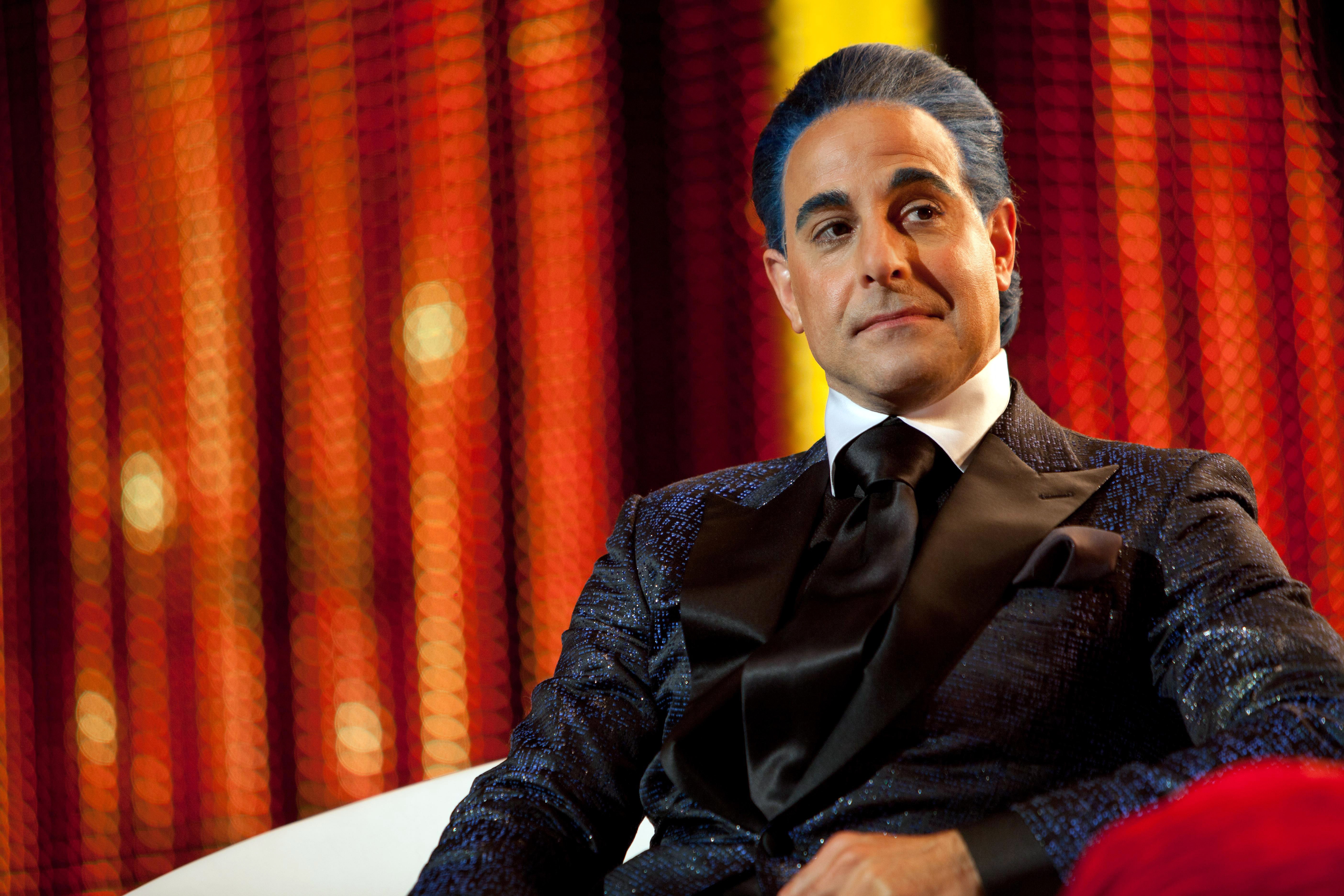 Stanley with slicked-bakc hair and in a formal, textured suit with a shiny tie, sitting in a chair, against a backdrop of patterned drapes