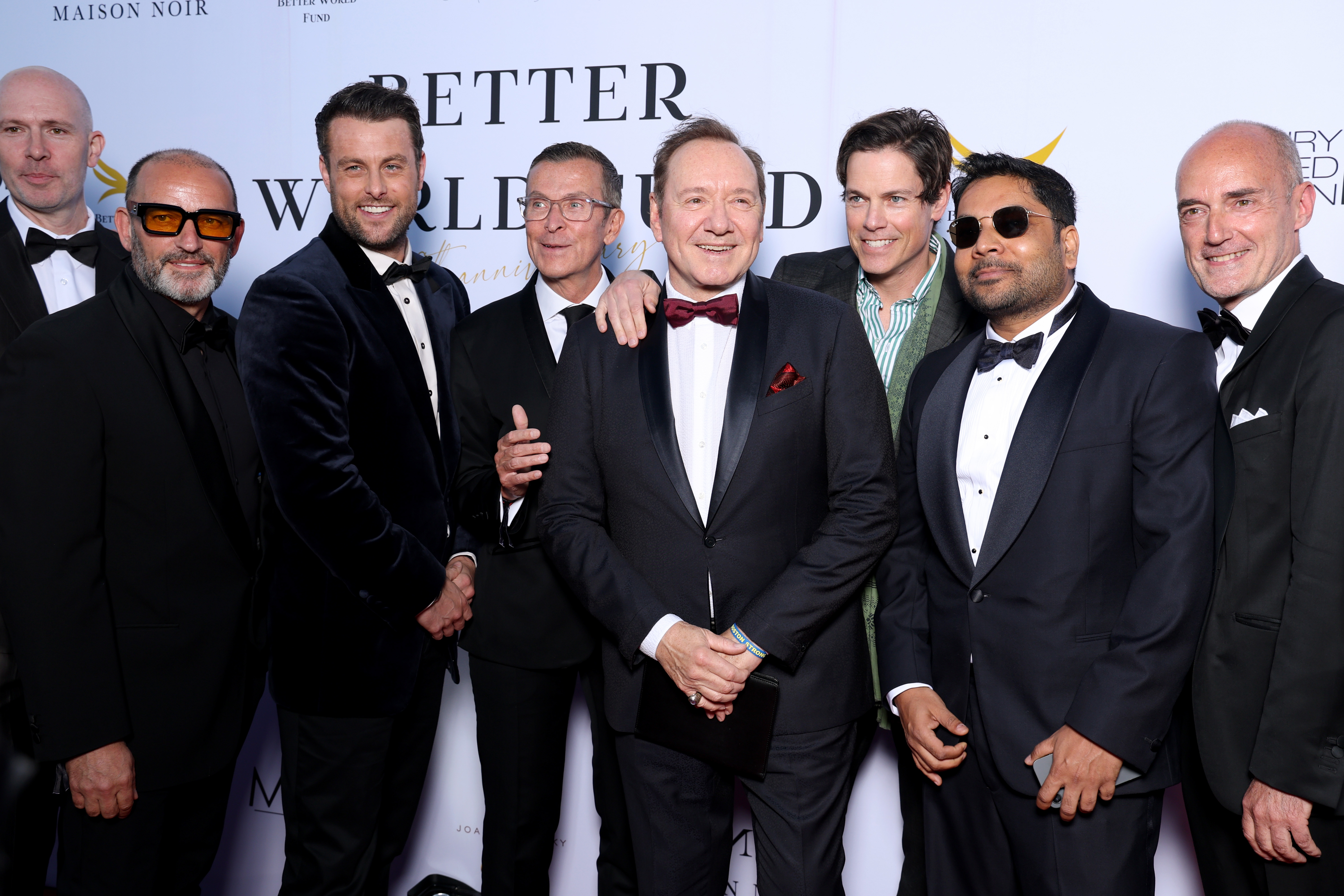Group of men in formal attire posing on a red carpet, some in tuxedos and bow ties, smiling for the camera