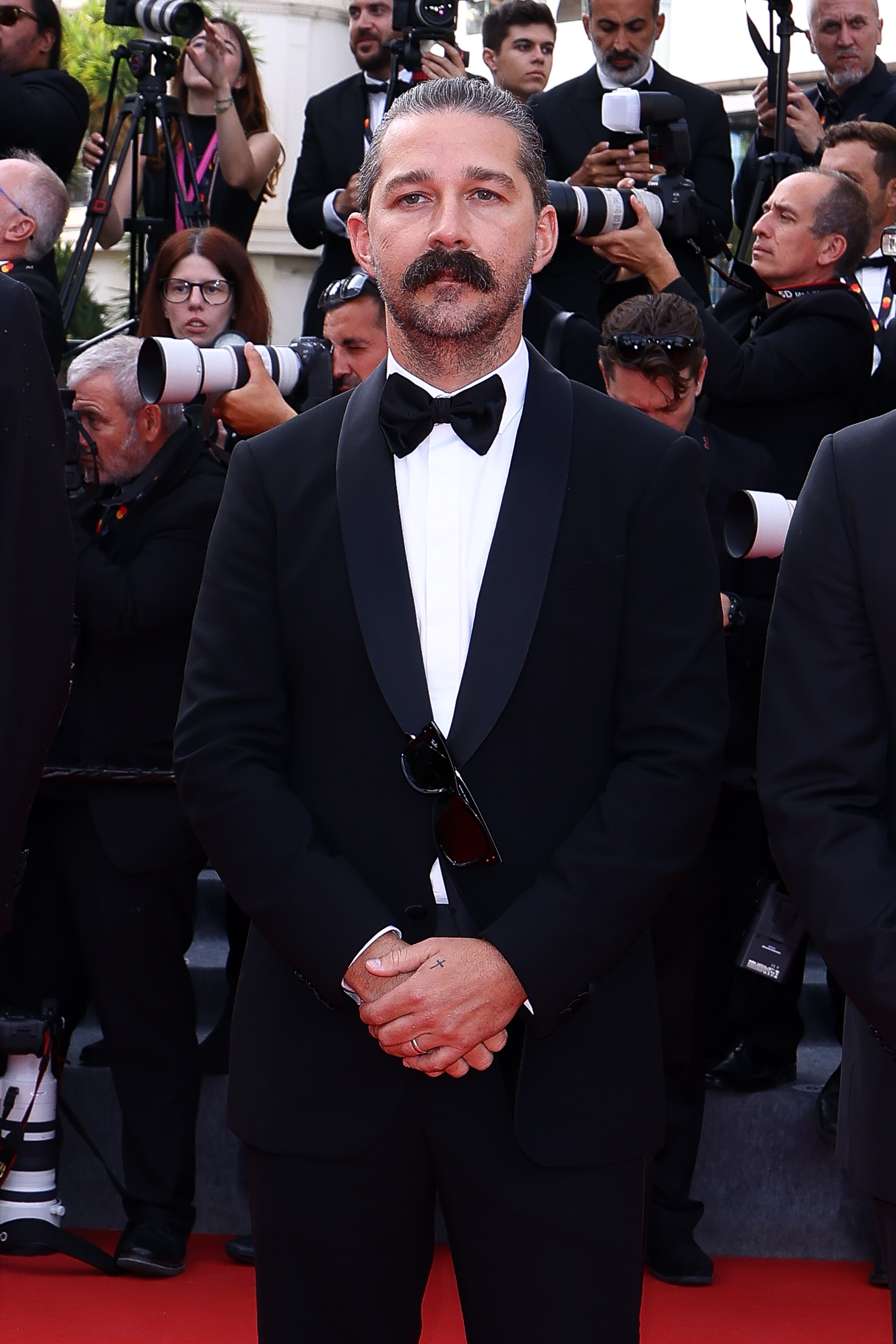A person in a classic tuxedo poses on a red carpet, with photographers in the background