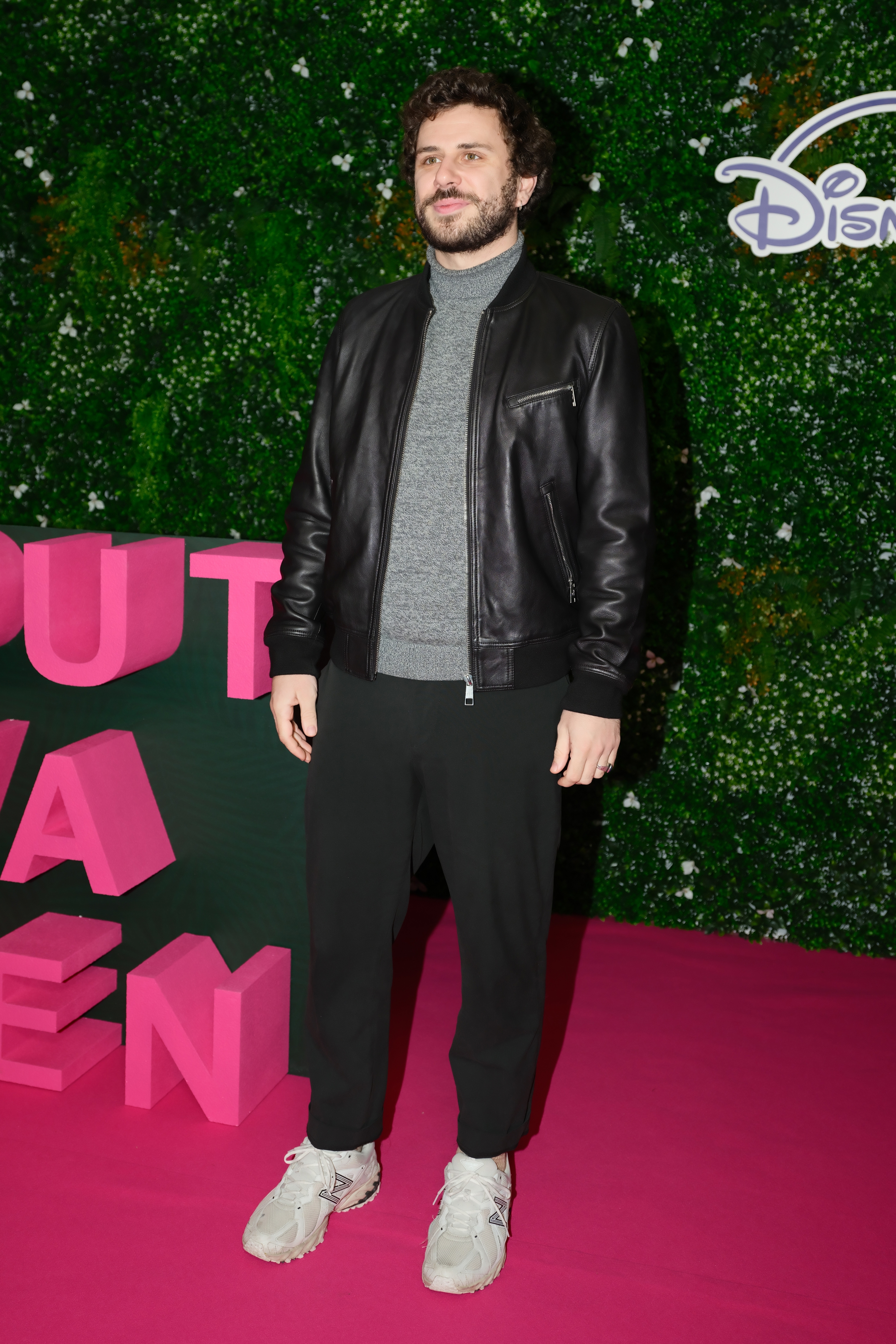 A person on a pink carpet wearing a black leather jacket, gray turtleneck, and black pants, standing in front of a greenery wall with Disney branding