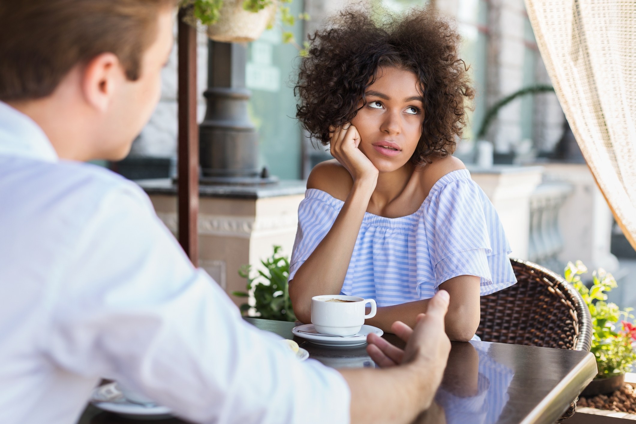 Two people sit at an outdoor café; one talks, while the other, appearing uninterested, rests their head on their hand