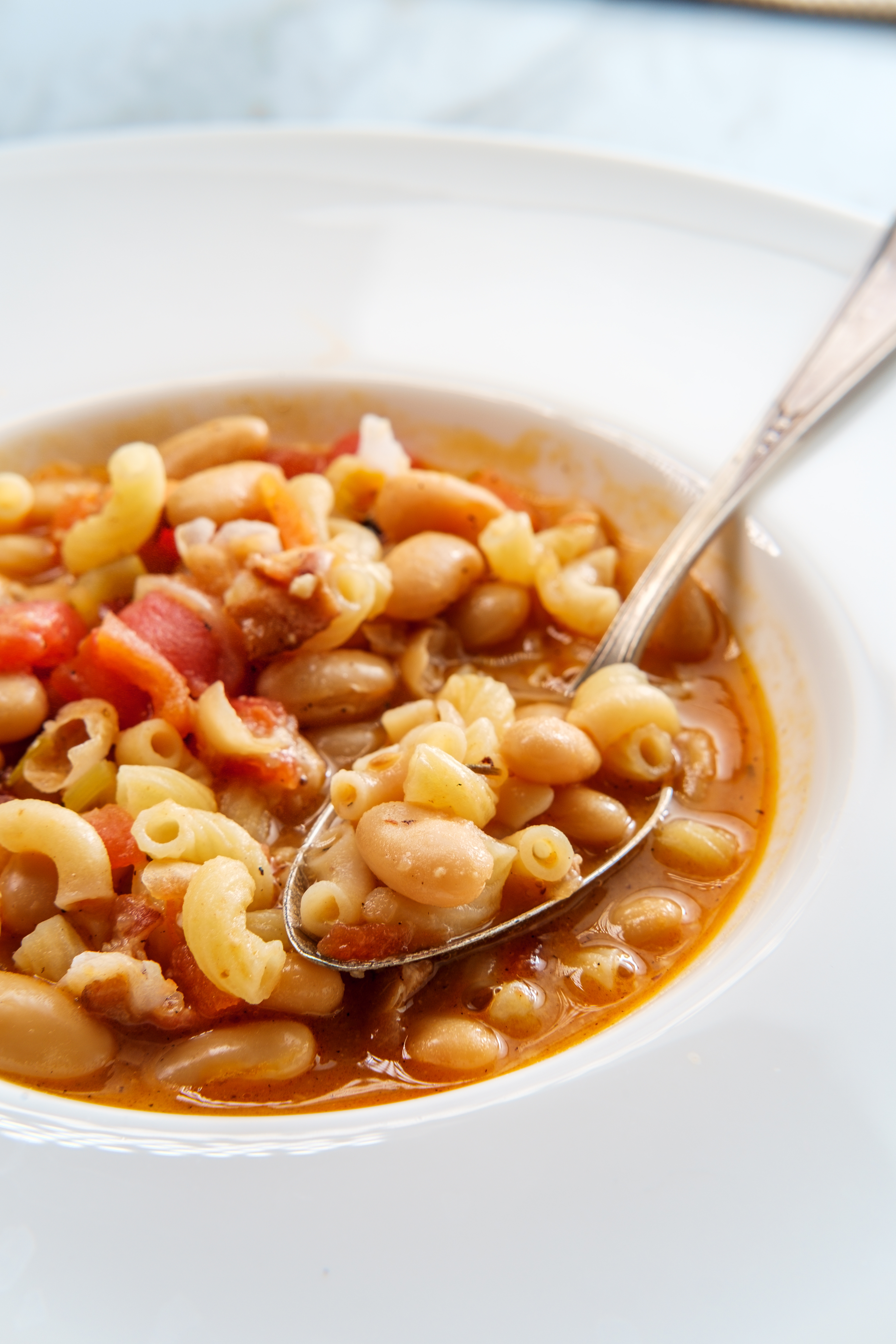 A close-up of a bowl of pasta e fagioli soup, featuring pasta, white beans, and tomatoes with broth, and a spoon resting on the bowl's edge