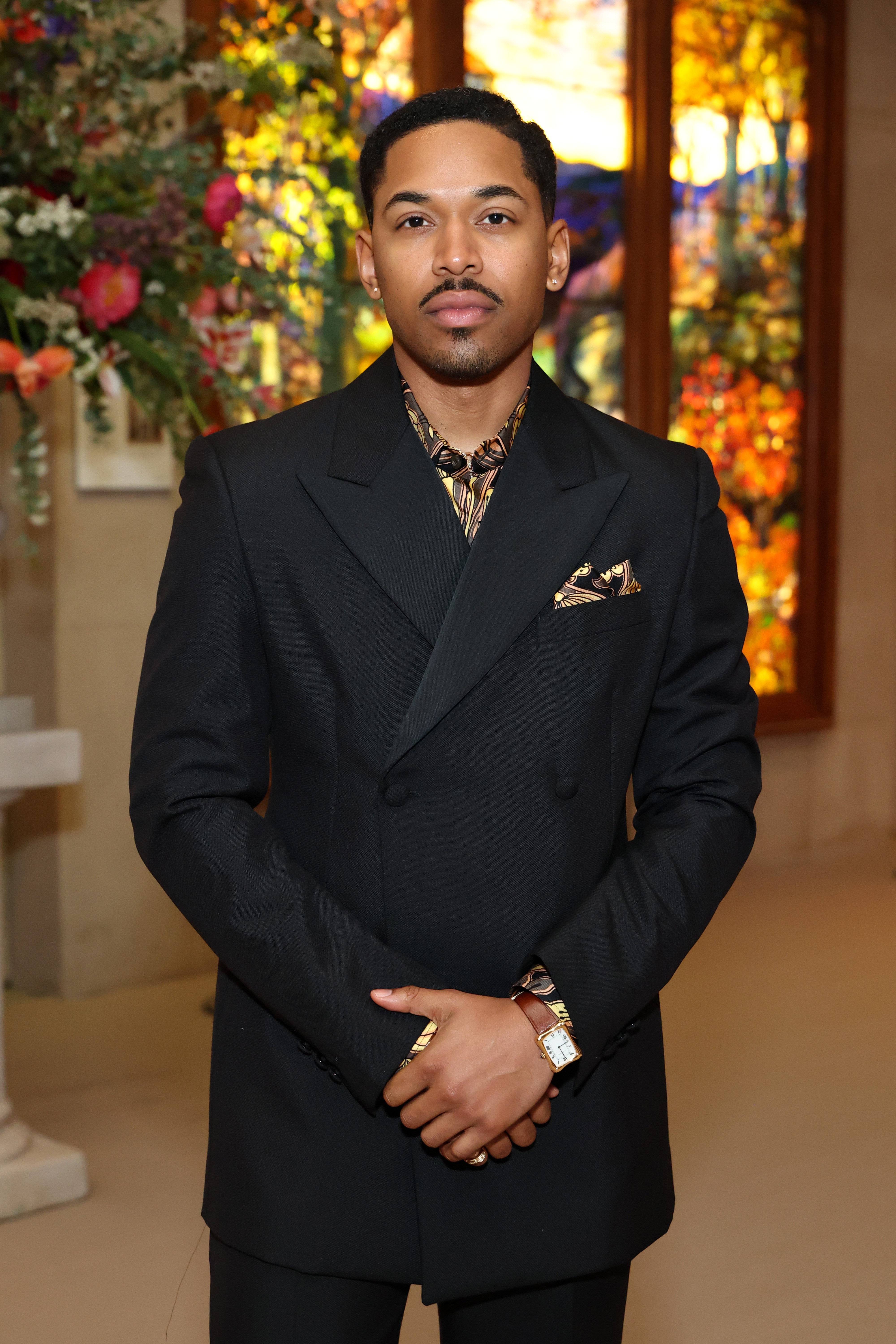 Kelvin in a stylish double-breasted suit with patterned shirt, standing inside near vibrant stained glass and floral decor