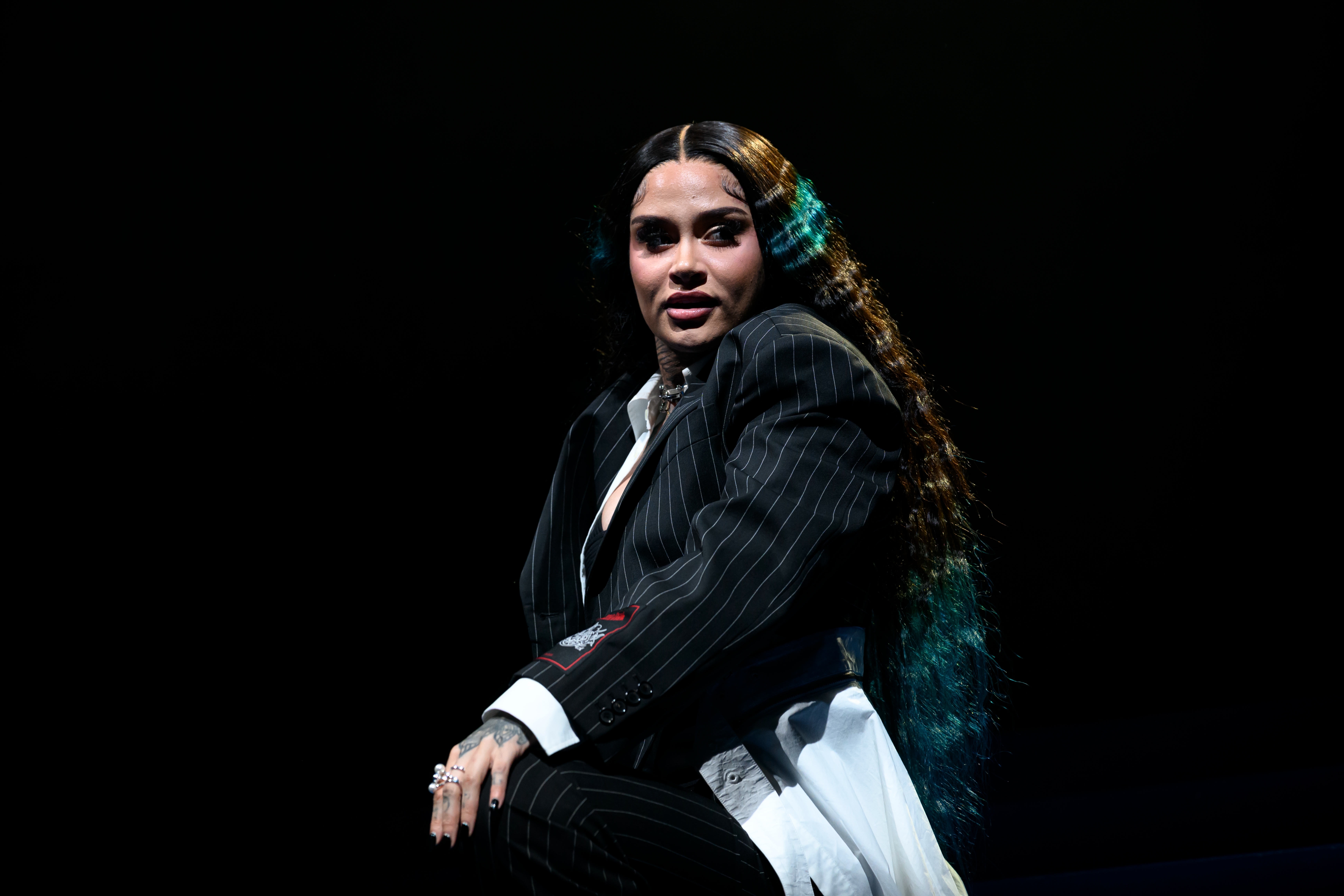 Kehlani in a pinstripe suit poses confidently on stage, with long wavy hair