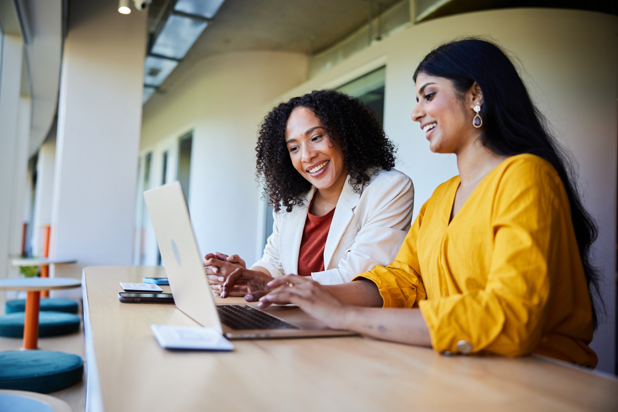 Two women smiling and working on a laptop at a long table outdoors, engaged in a collaborative discussion
