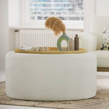 Round achromatic ottoman holds a tray with a vase of dried flowers, 2 bottles, and a book, placed successful a modern surviving room