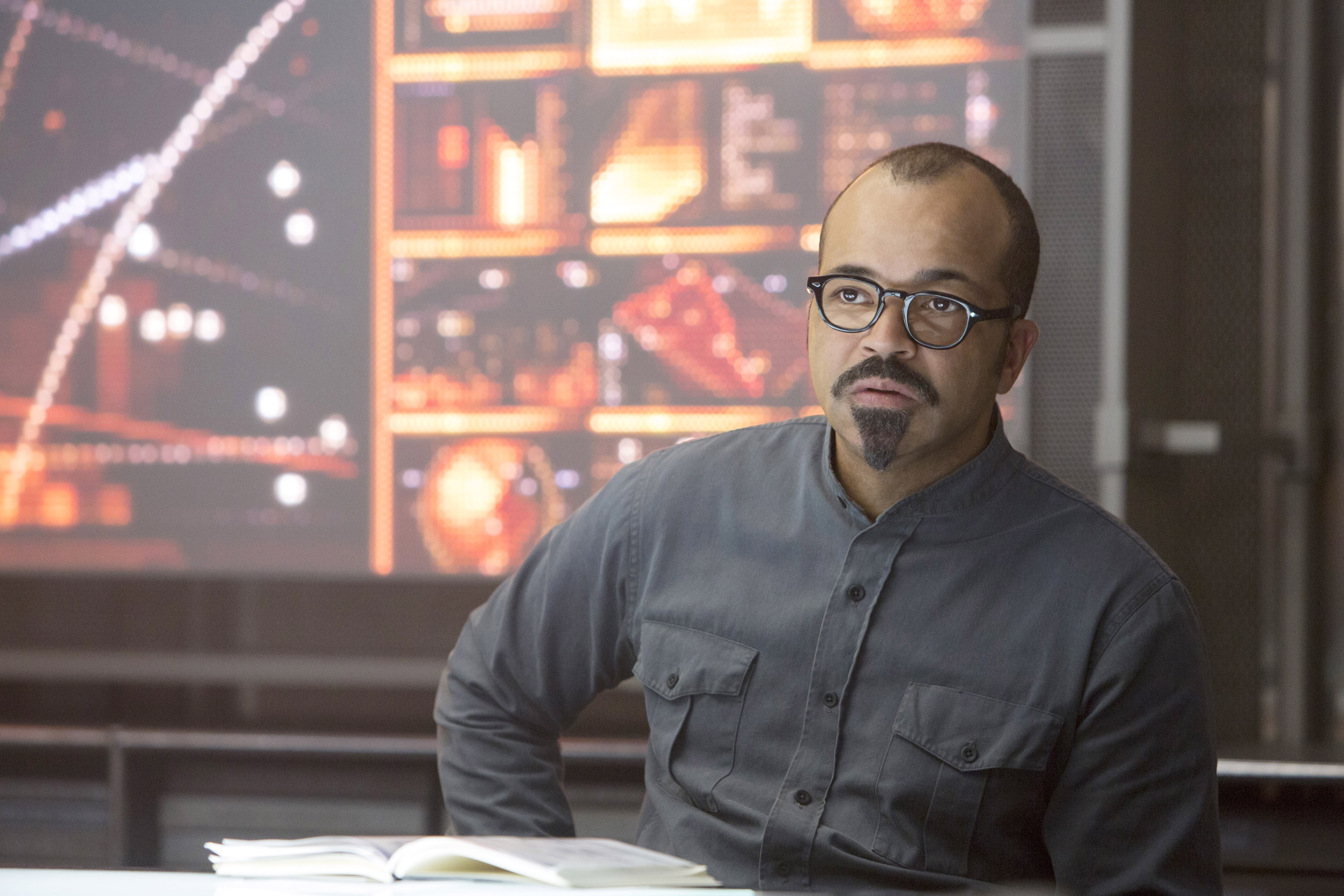 Jeffrey in glasses and a buttoned shirt sitting at a desk, with abstract visuals displayed on a screen behind him