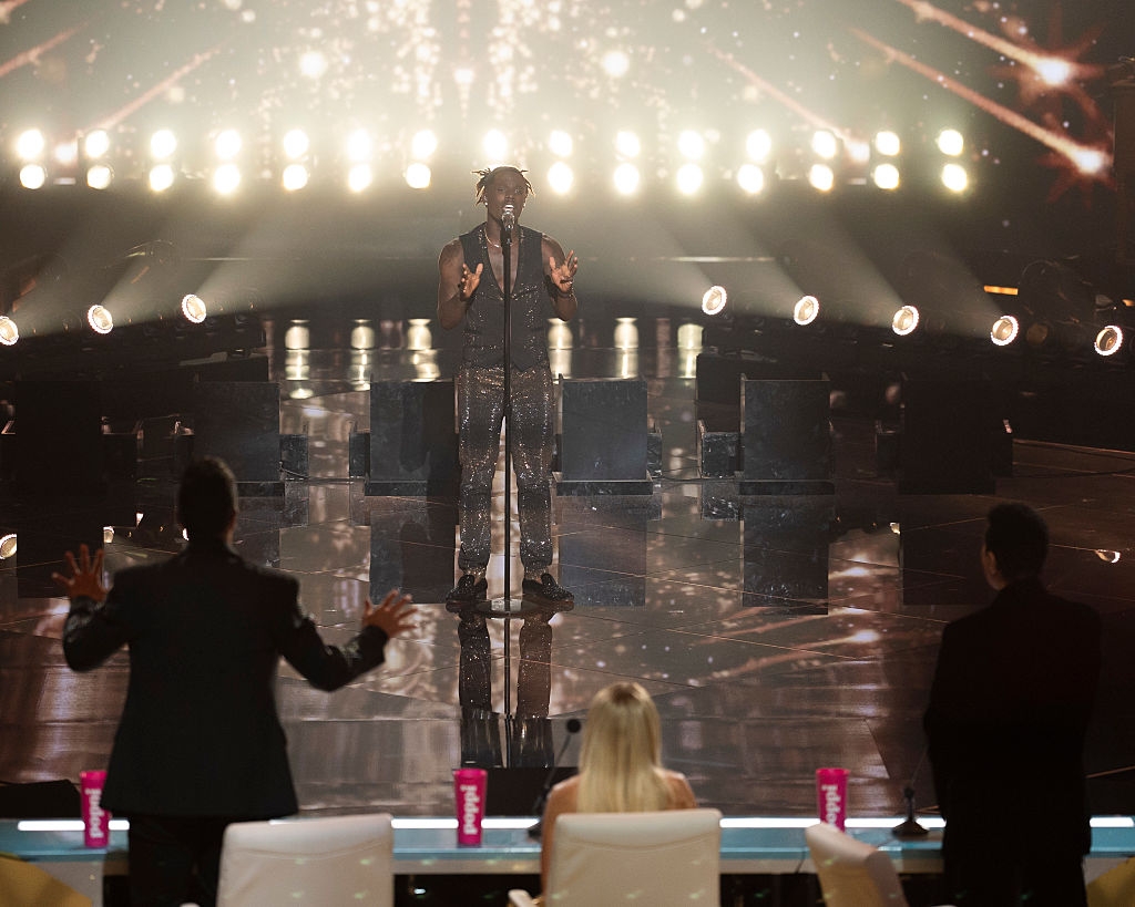 Performer on stage during a talent show with judges in the foreground and bright stage lights in the background