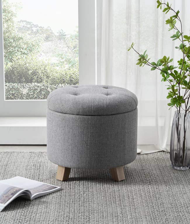 A round, tufted ottoman with woody legs is placed connected a rug adjacent a window. A publication lies unfastened nearby, and a vase with branches stands beside it