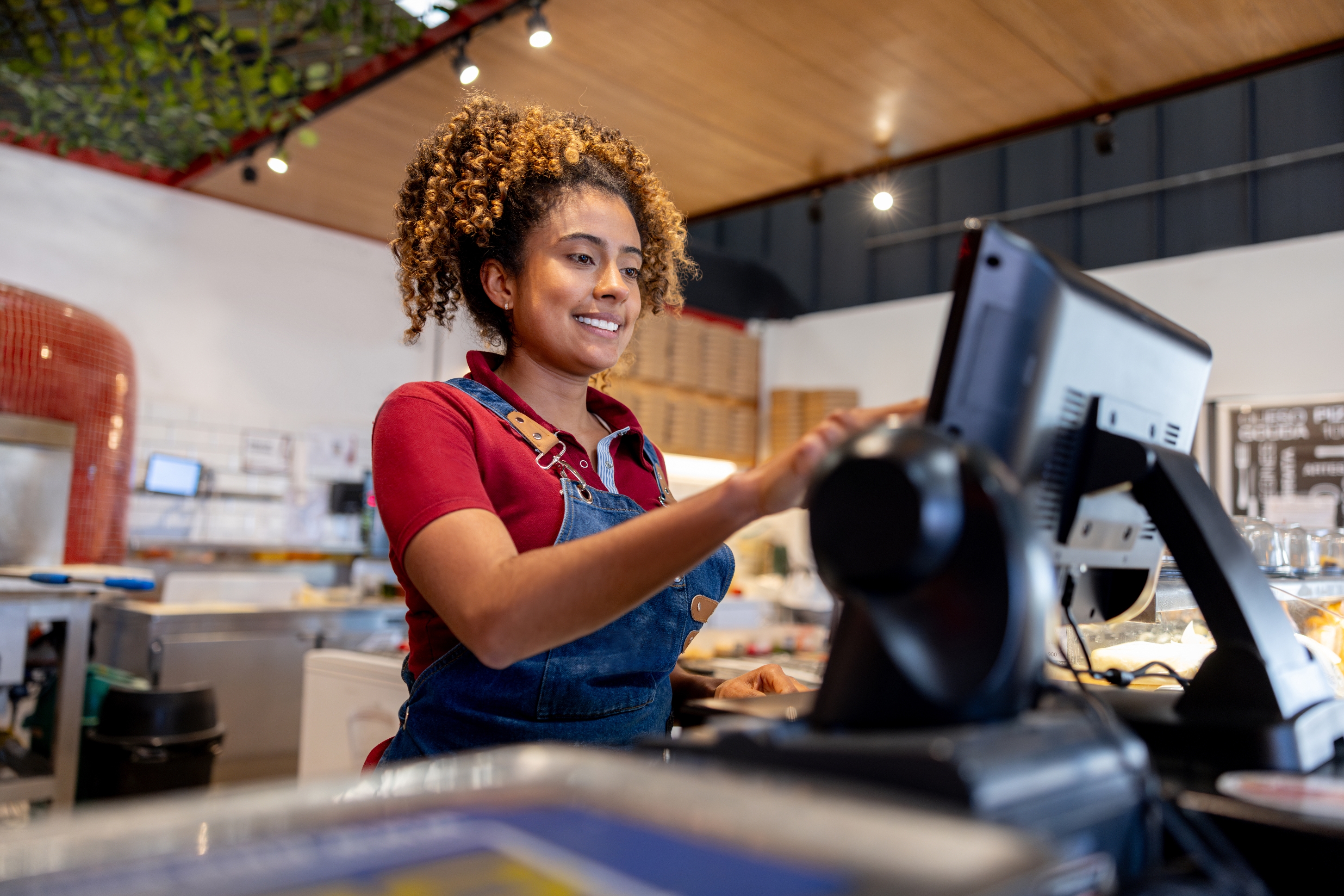 Person in a café, wearing a denim apron, smiles while working on a computer at the counter