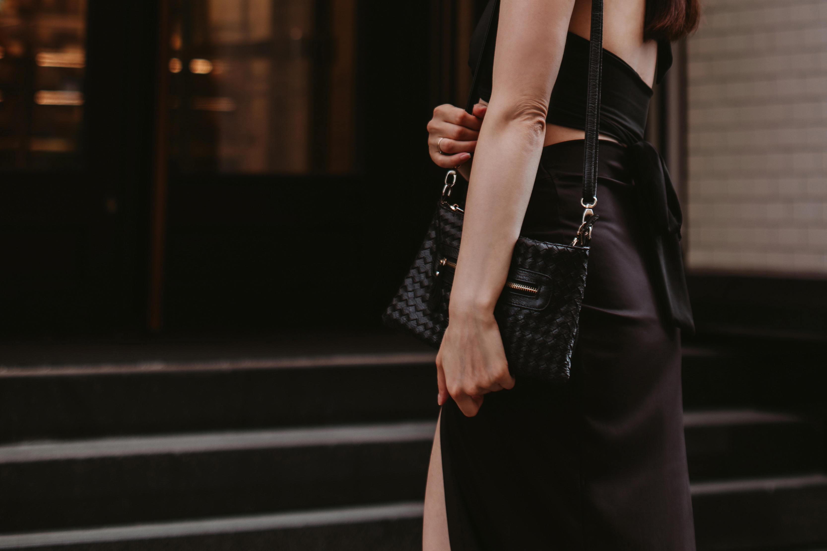 A person wearing an elegant black outfit holds a textured handbag, standing on steps outside, suggesting a sophisticated setting