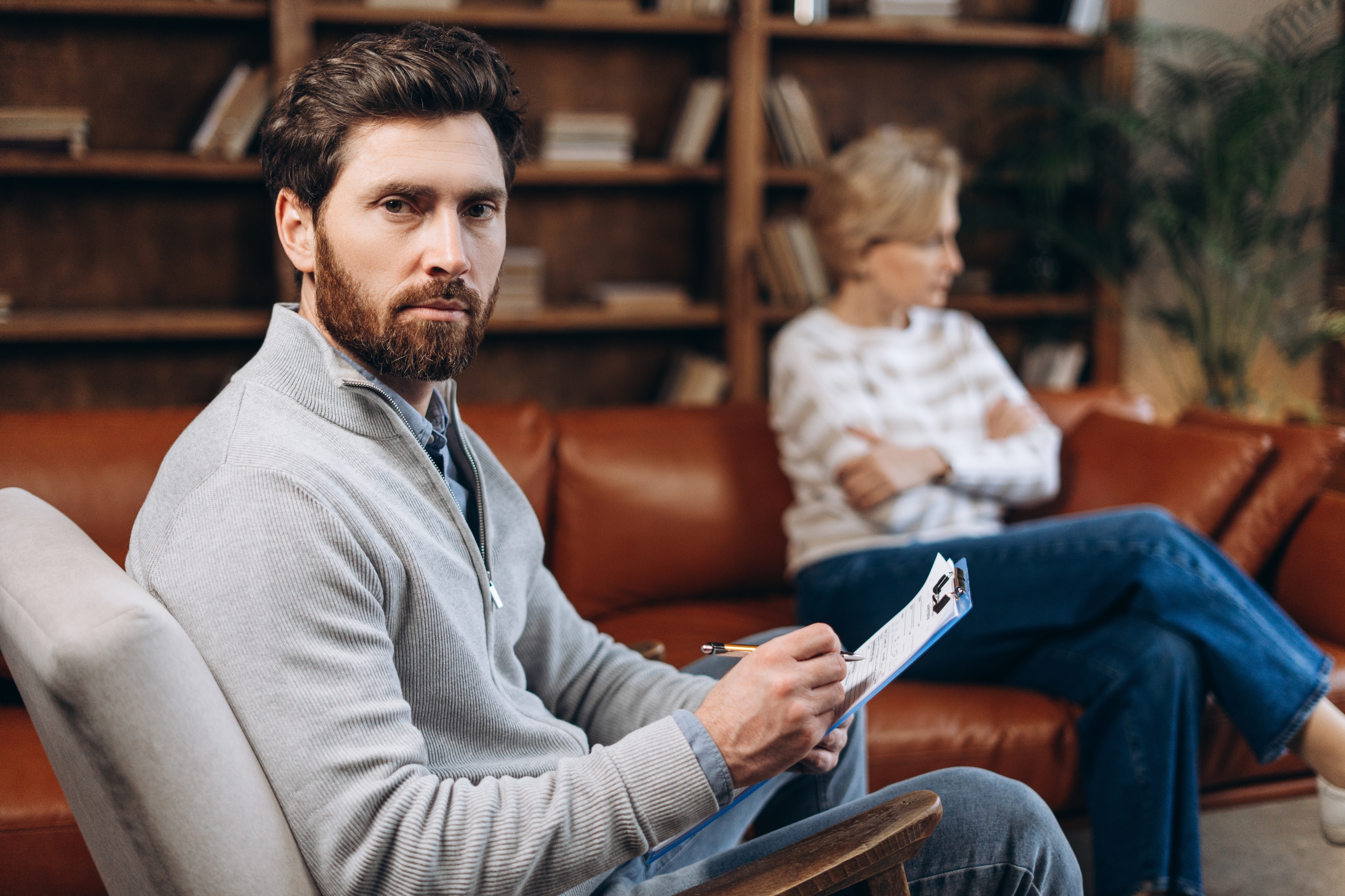 Man with a serious expression holds a clipboard, seated on a couch; woman in the background looks away, suggesting tension or disagreement