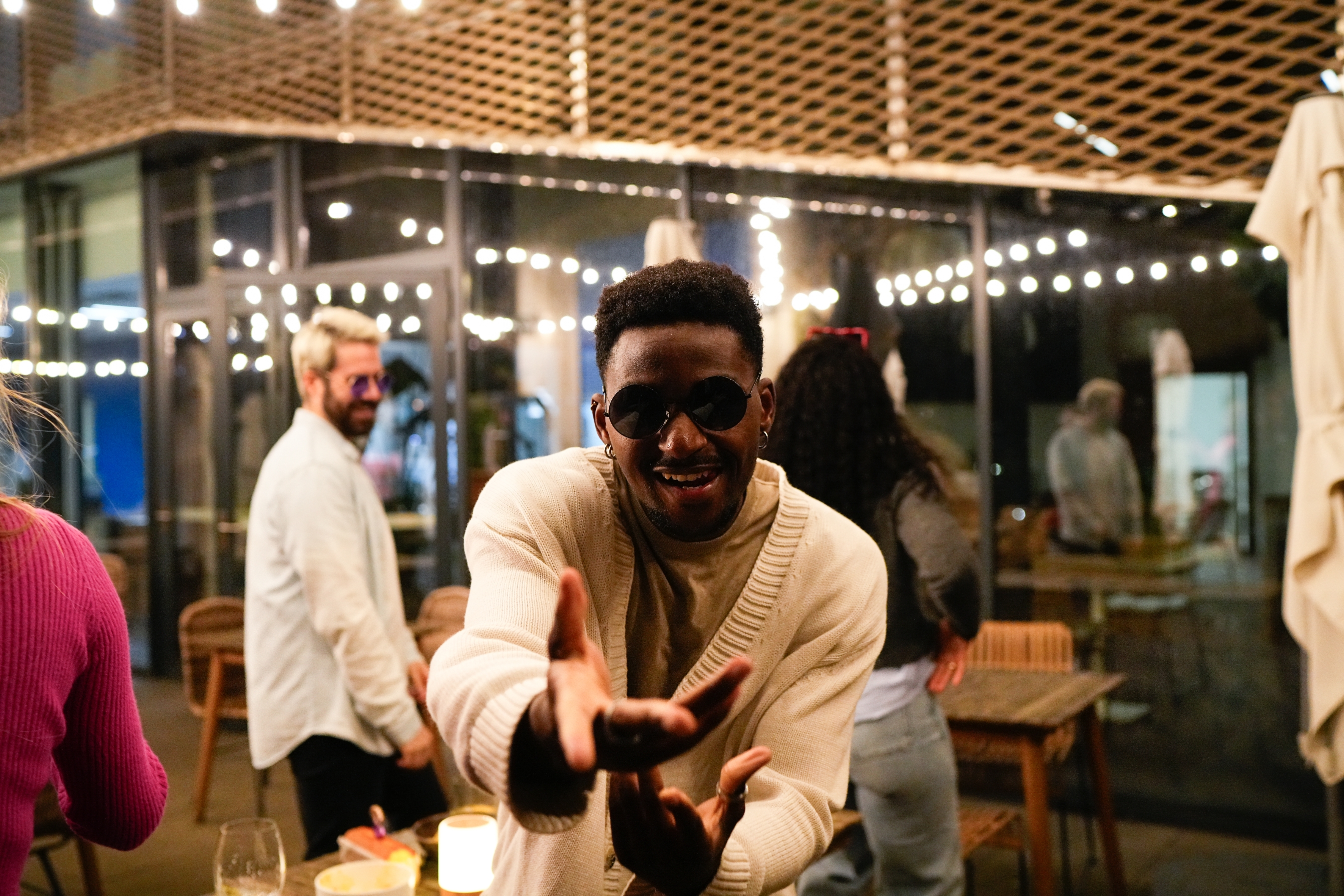 Man in cozy sweater and sunglasses poses playfully, arms reaching out, at a lively social gathering