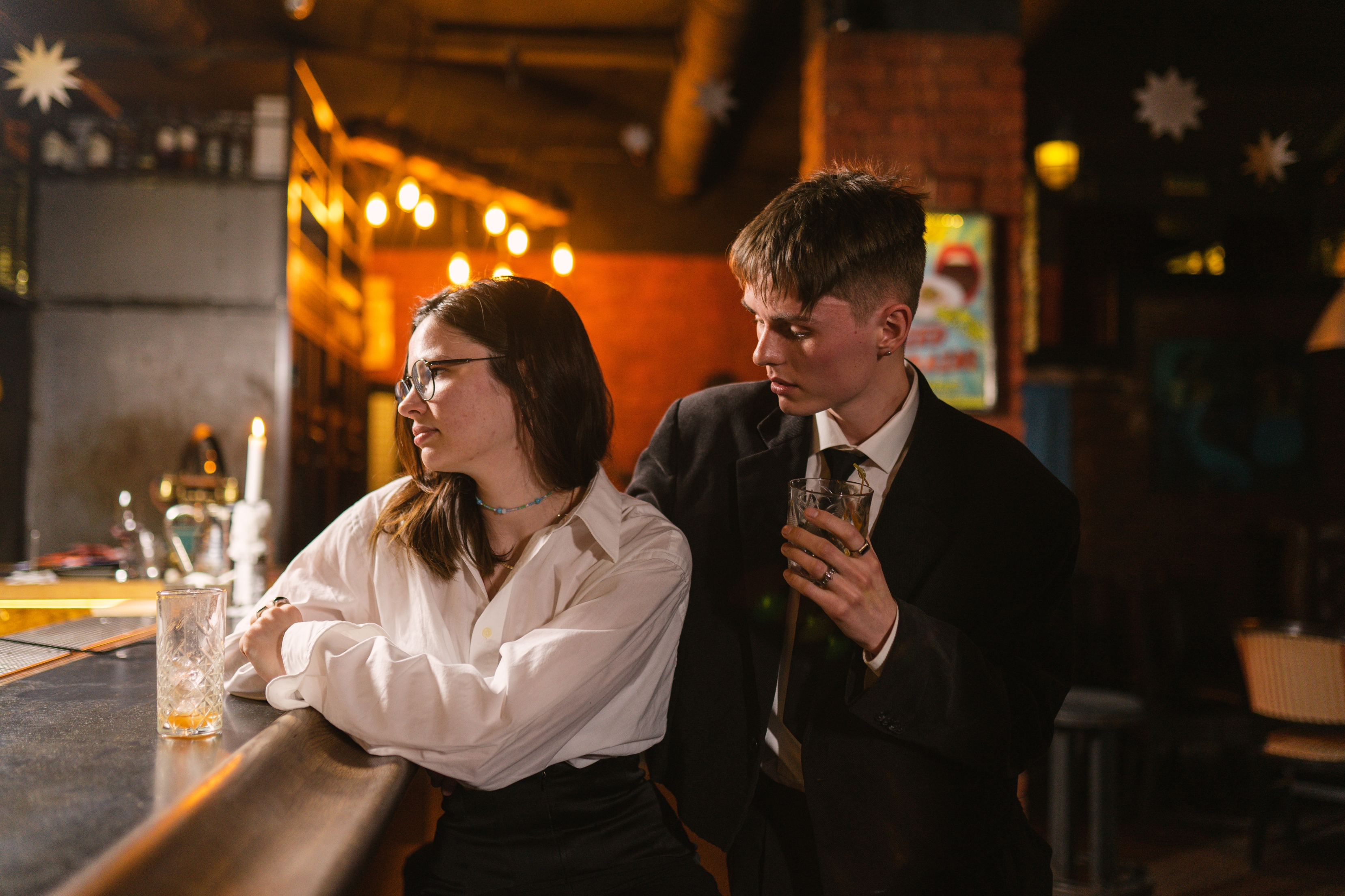 Two adults in a bar; one leans on the counter in a white shirt, looking away. The other, in a suit, holds a drink, gazing at the first person