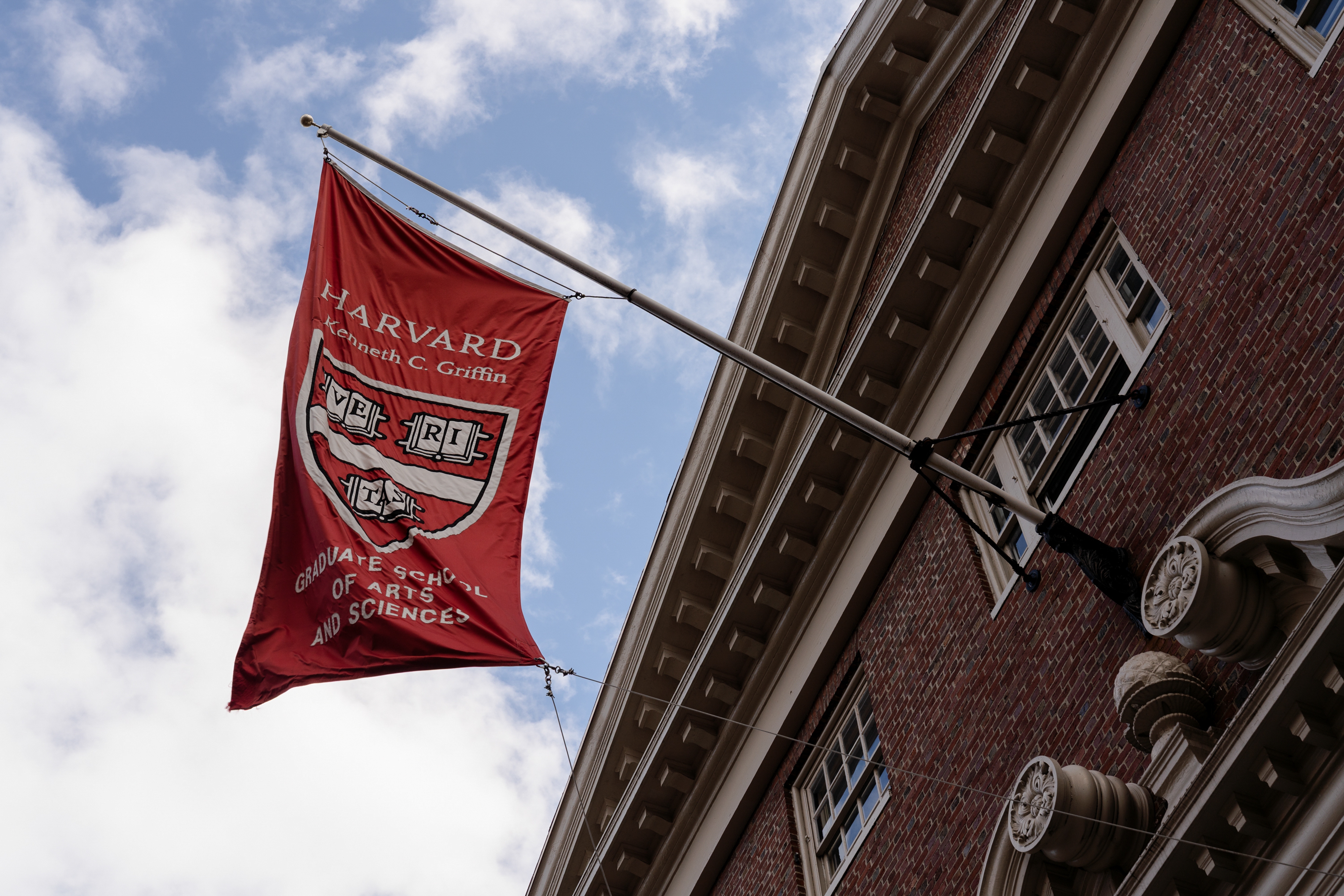 A Harvard University flag with the motto &quot;Veritas&quot; hangs from a building. The flag includes the Graduate School of Arts and Sciences text