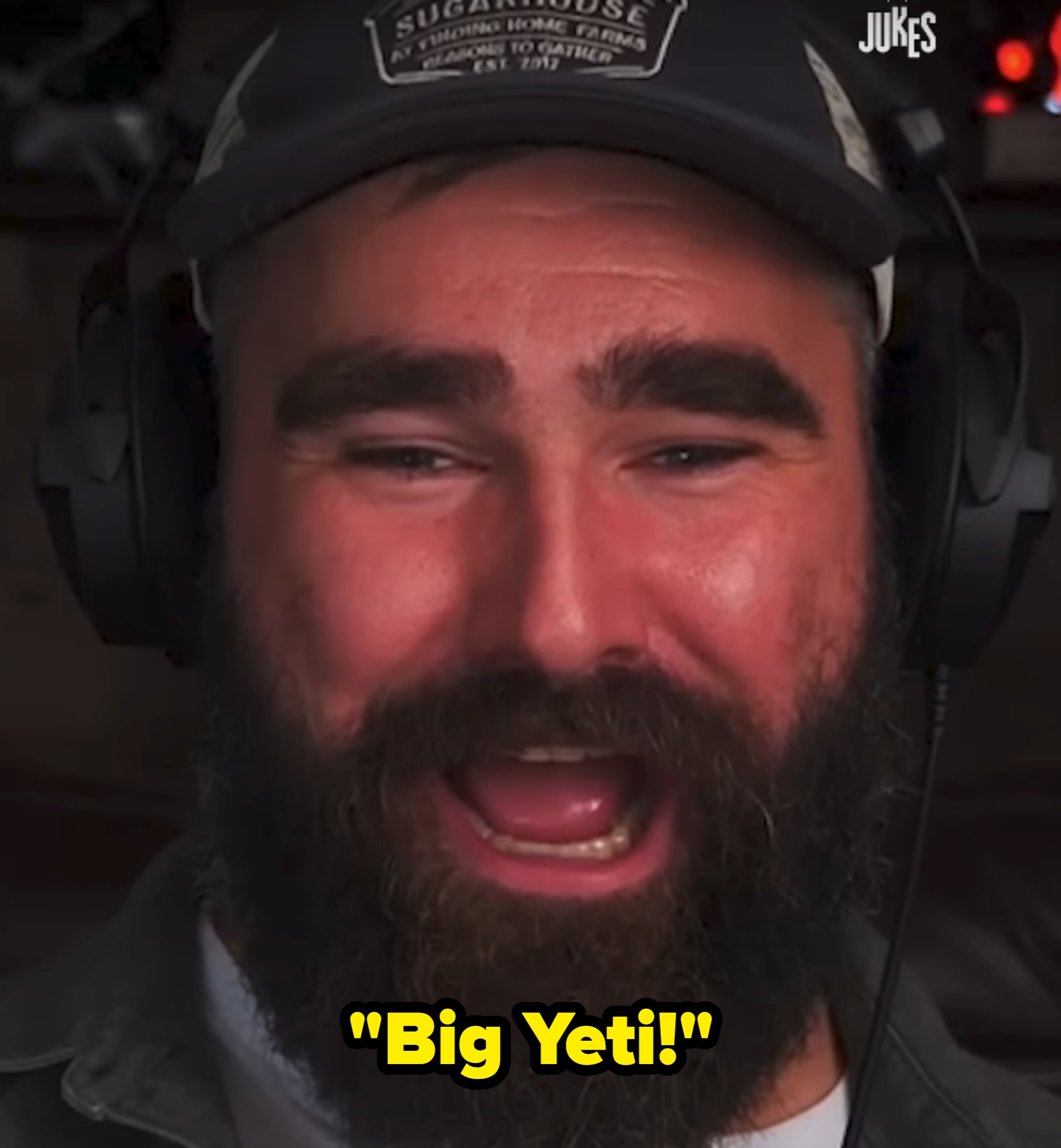 A man with a beard and headphones laughs, wearing a cap and jacket