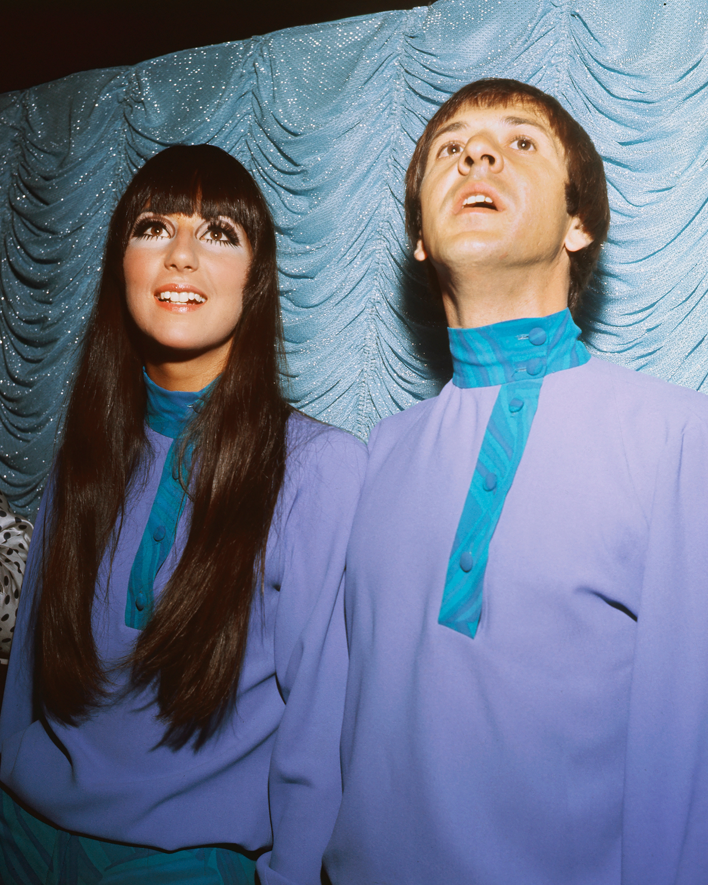 Two individuals in vintage outfits with long sleeves and unique collars stand against a textured backdrop, gazing upward