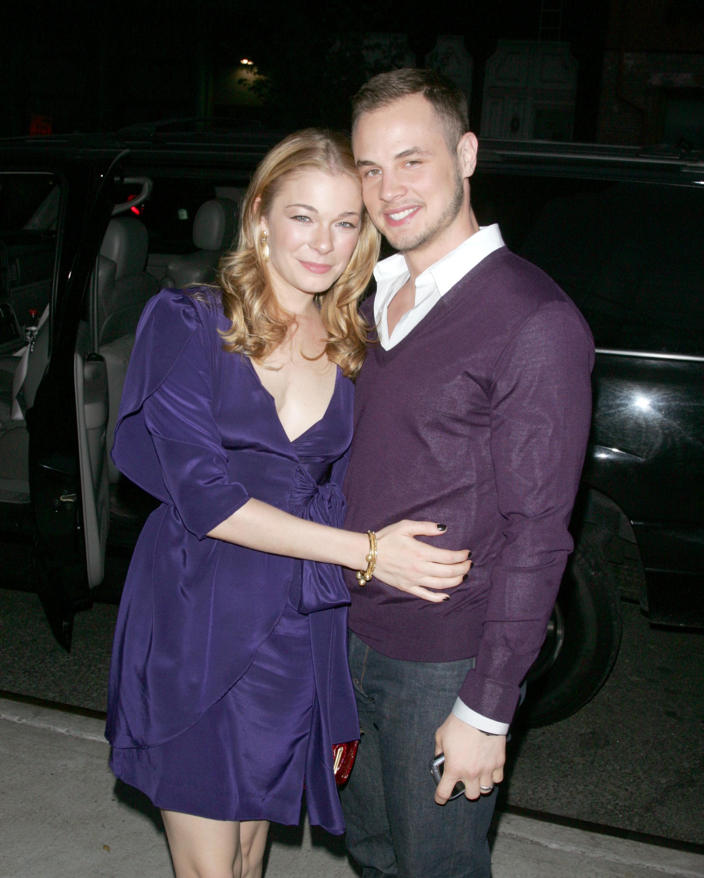 Two celebrities smiling and posing outside, with one wearing a stylish dress and the other in a casual V-neck sweater, standing near a vehicle