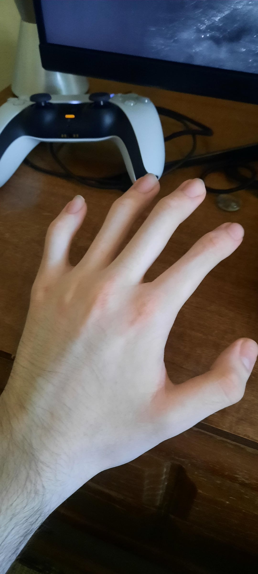 A person's hand rests on a wooden desk near a video game controller and a computer monitor