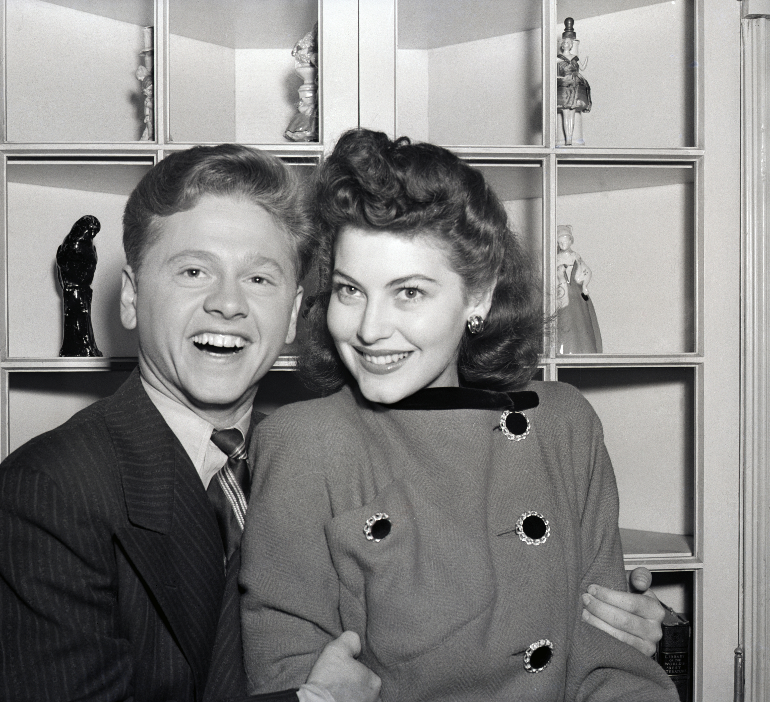 A smiling couple from the mid-20th century, dressed in formal attire with classic hairstyles, posing indoors in front of a shelf with figurines