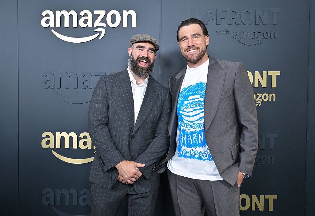 Two men smiling at an Amazon event. One wears a suit and cap, the other a suit with a graphic T-shirt. "UPFRONT with Amazon" is seen in the background