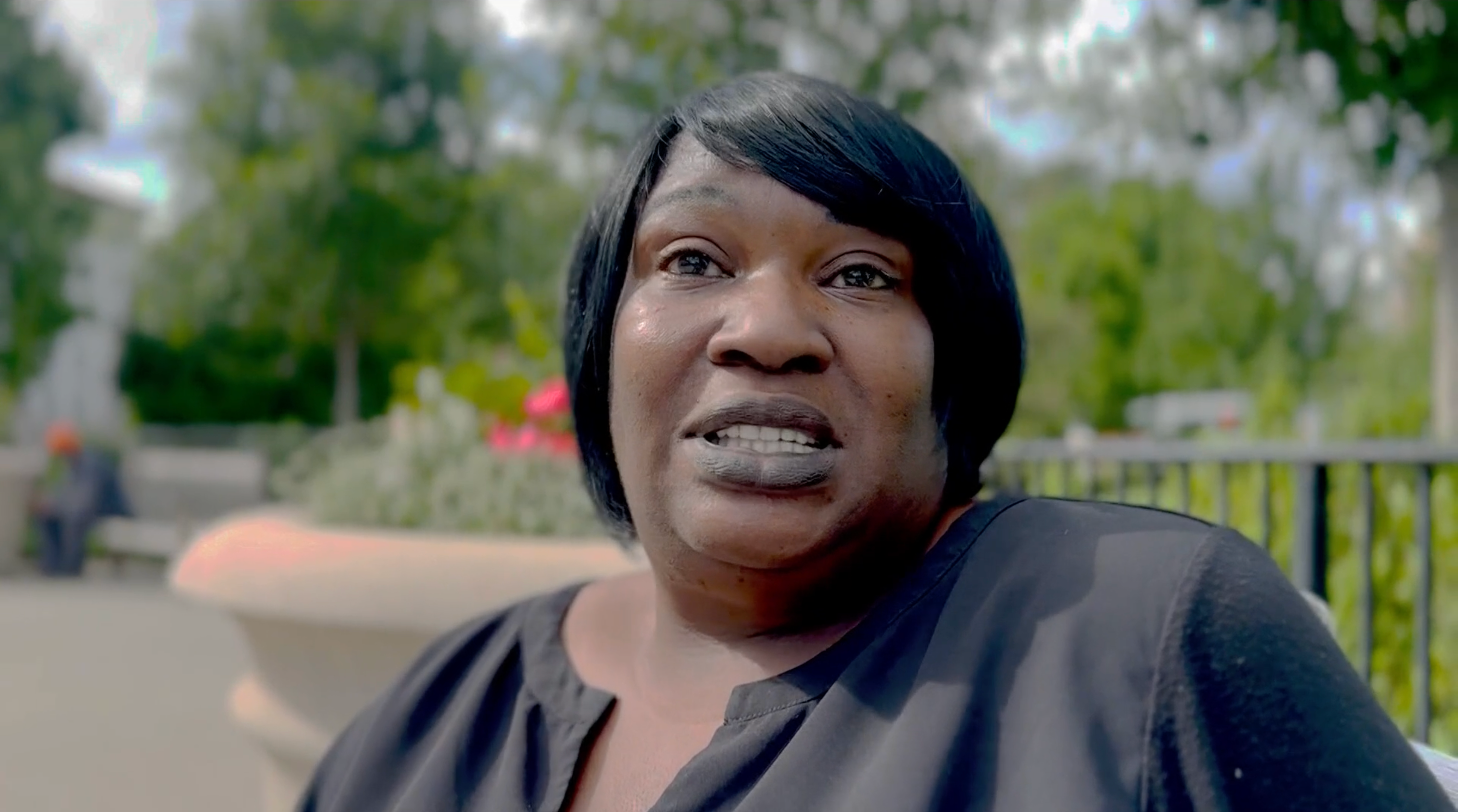 A person sits outdoors, speaking in a garden setting with trees and flowers in the background