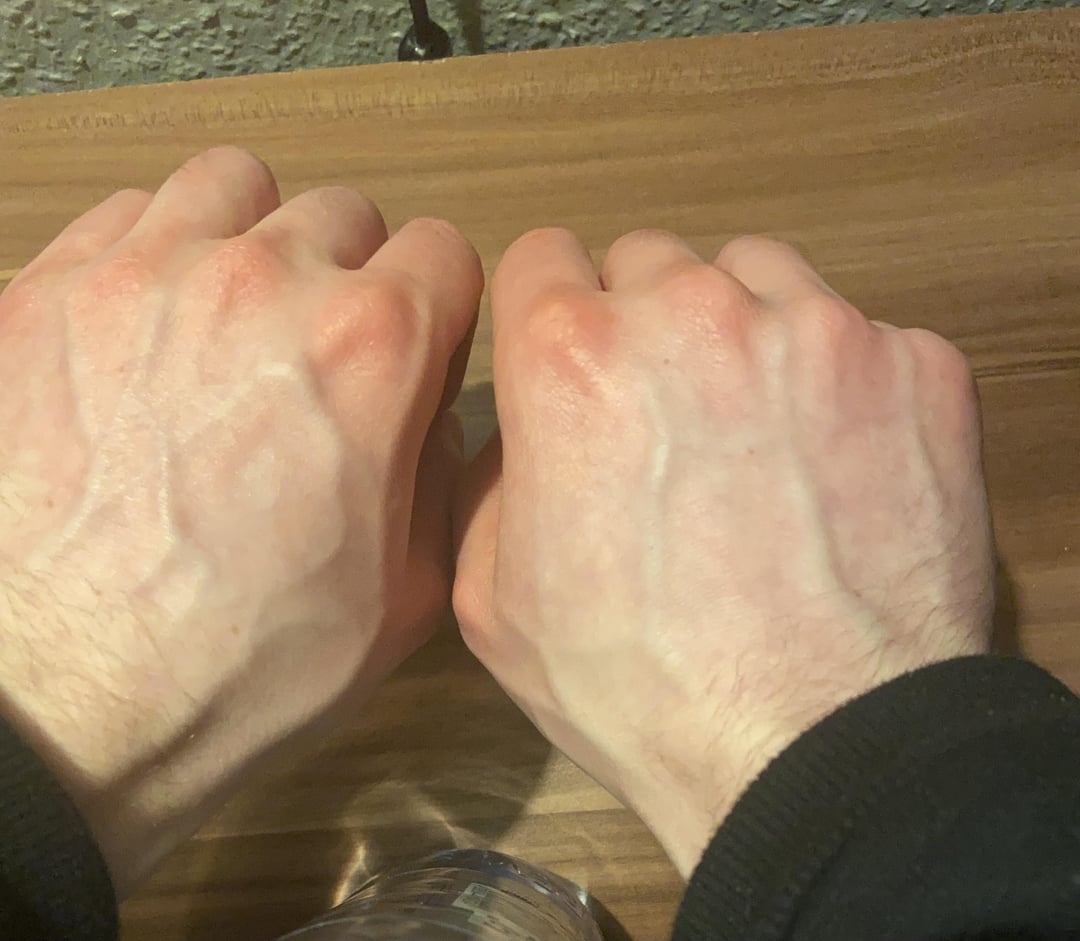 Two hands resting on a wooden surface, veins visibly prominent. A water bottle cap is partially visible at the bottom of the image