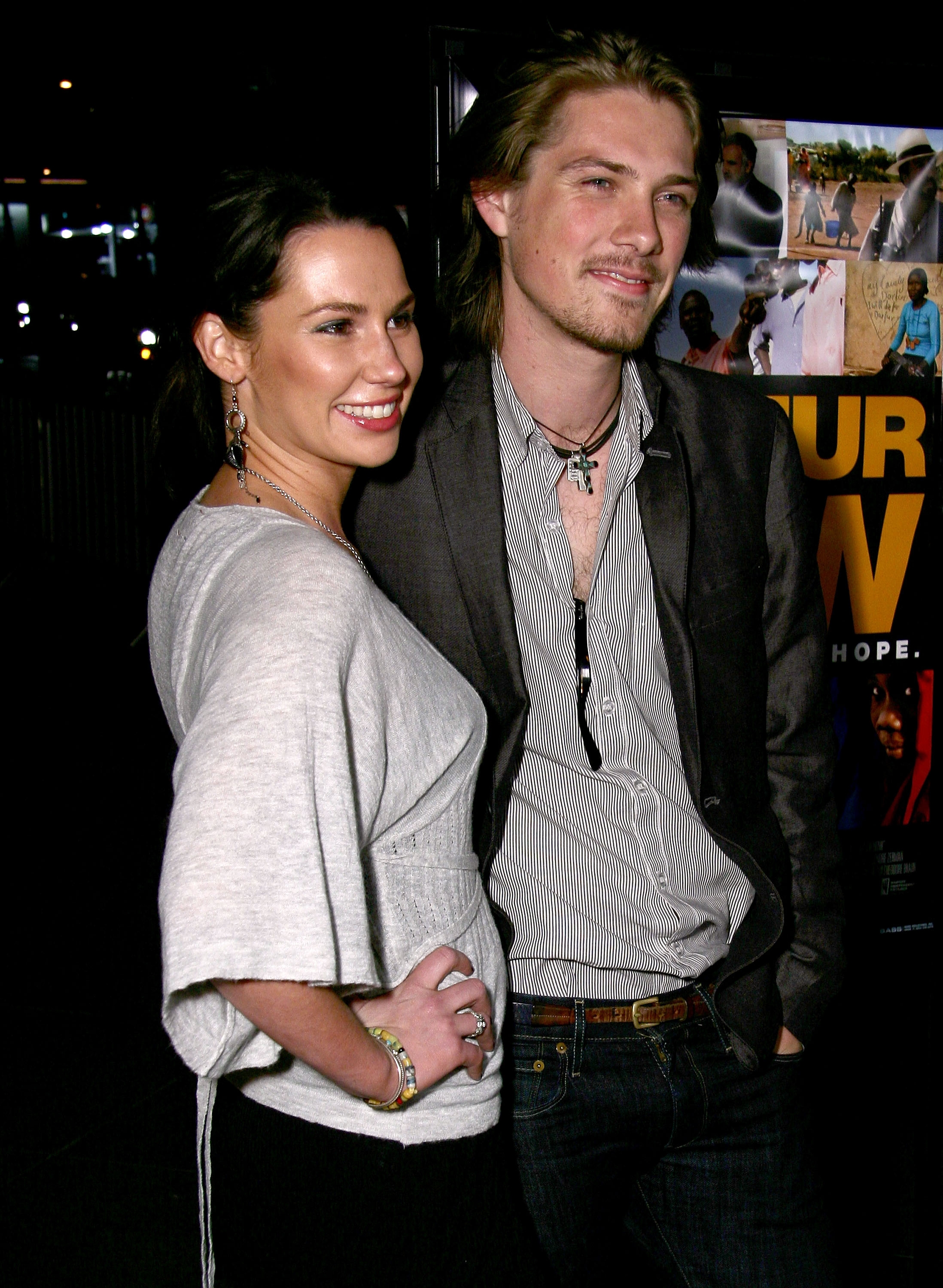 Two individuals pose together at an event. The man wears a casual blazer over a striped shirt, and the woman is in a stylish top
