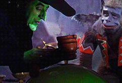 A witch with green skin holds a cup while a flying monkey watches. They are in a mysterious setting with a glowing orb