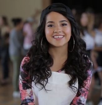 Persona sonriente con cabello rizado, gorra hacia atrás y aretes grandes. Lleva una prenda con estampado floral. Fondo desenfocado
