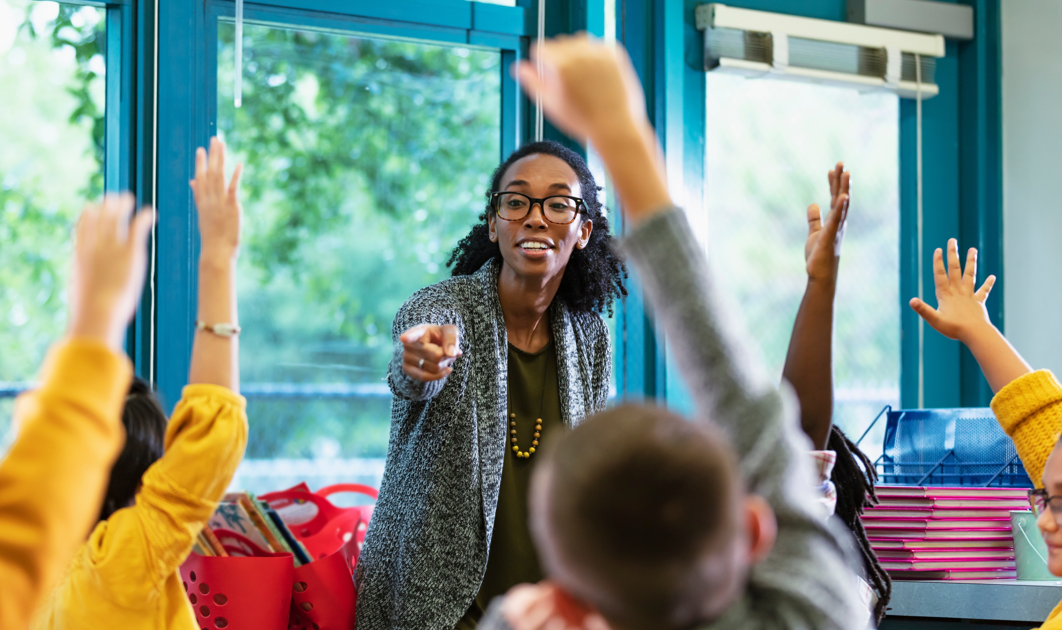 Teacher engaging with students, pointing to one with raised hands in a lively classroom