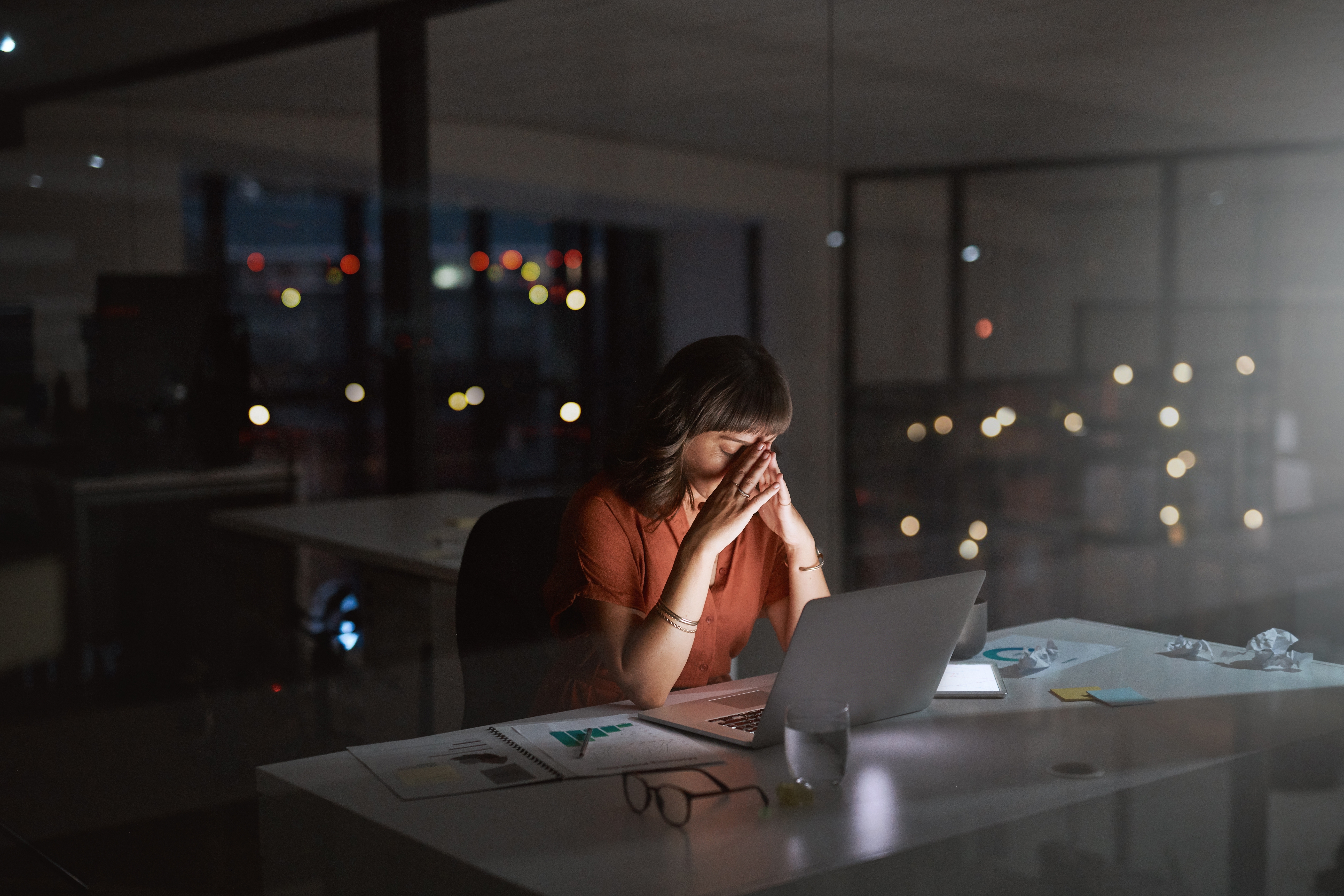 Person in an office at night, sitting at a desk with a laptop, holding their head in their hands, appearing stressed
