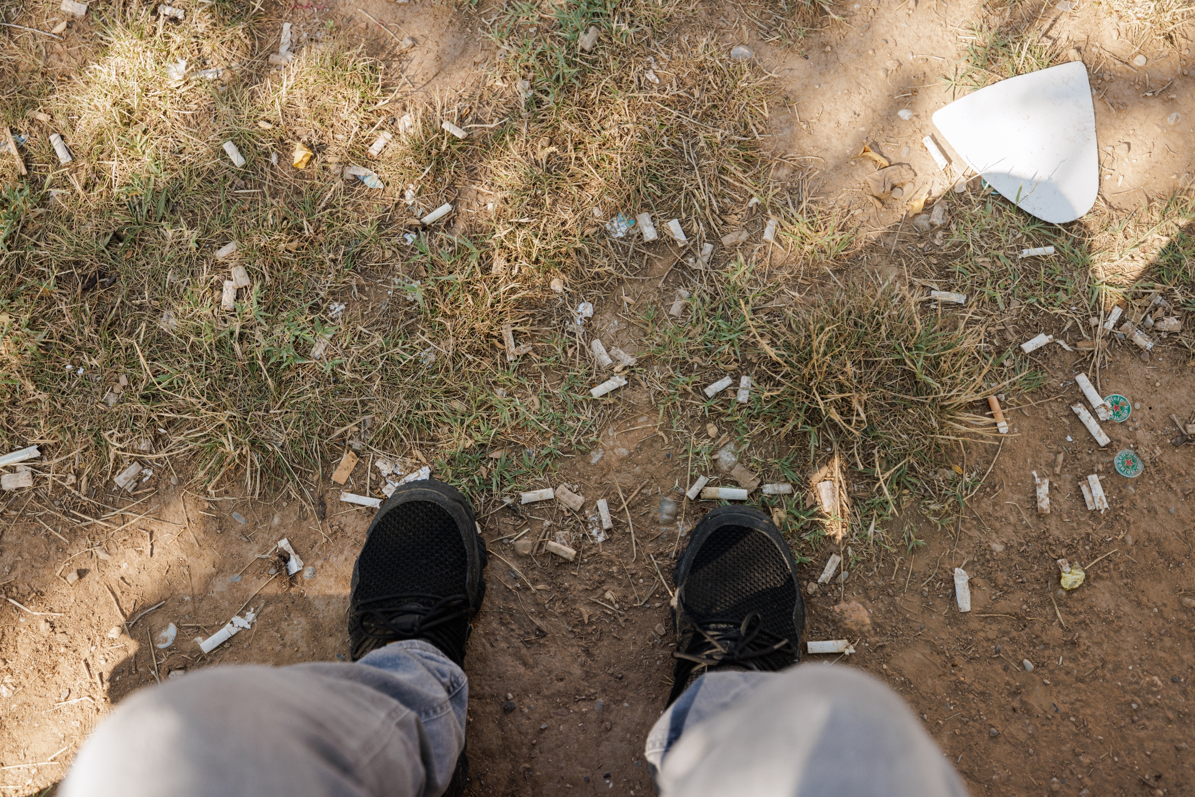 Feet in sneakers standing on a dirt ground scattered with cigarette butts and litter