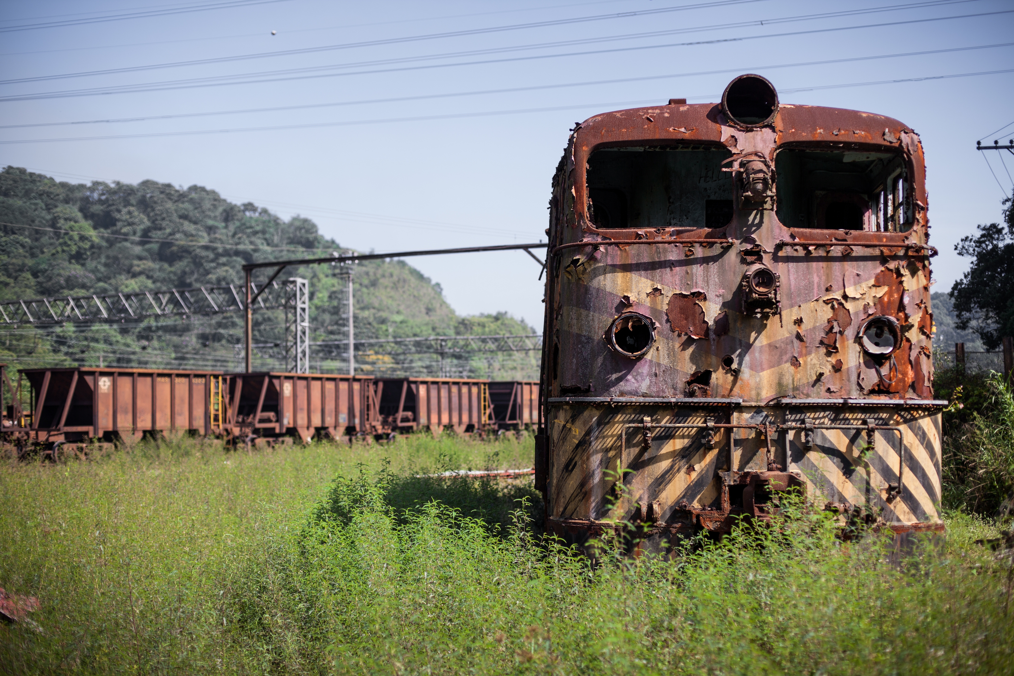 Rusty train engine and cargo cars sit abandoned on overgrown tracks, with a background of hills and power lines