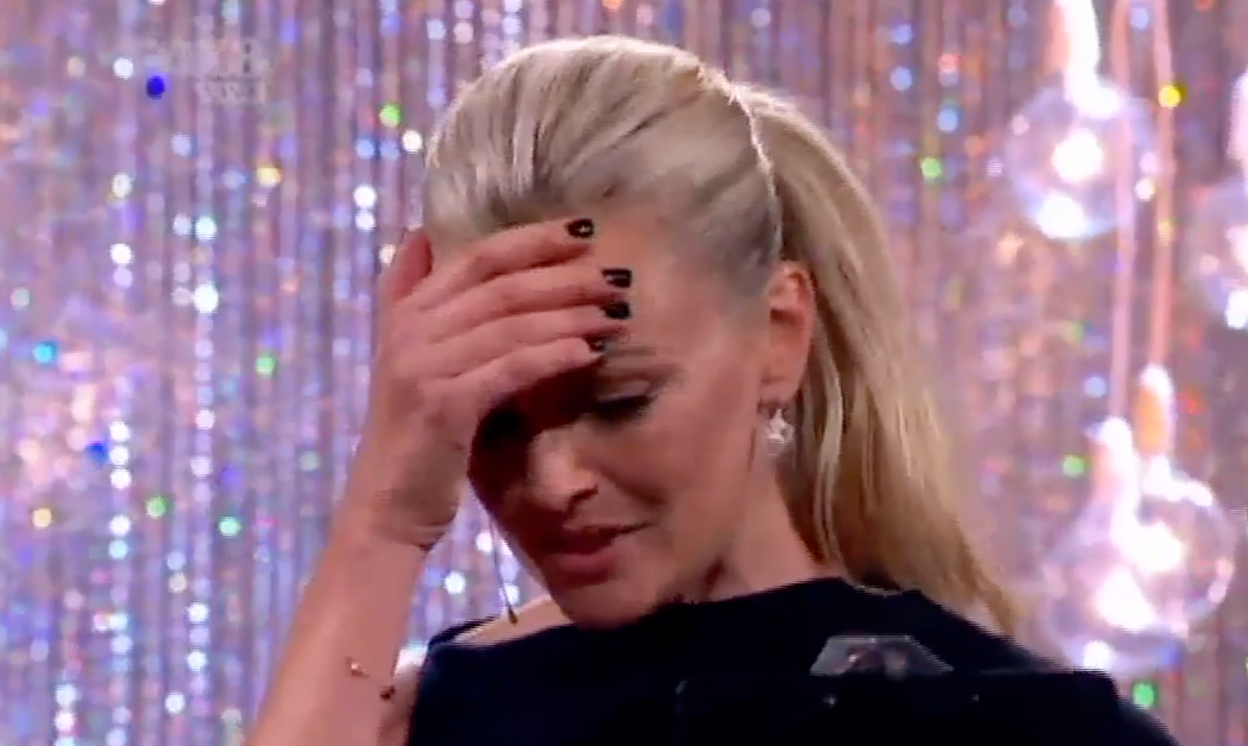 Person on stage with a ponytail, holding their forehead with one hand, possibly emotional or contemplative against a sparkly backdrop